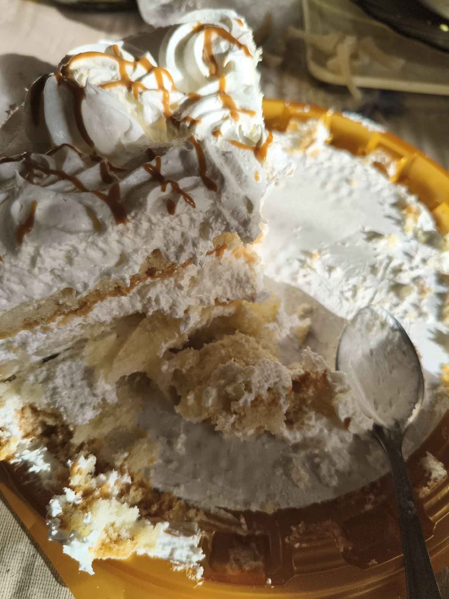 A slice of partially eaten cake with white frosting and caramel drizzle is on a yellow plastic plate. A spoon rests in the remaining cake. The scene is brightly lit with natural light, casting shadows. The background is out of focus, showing hints of a table and other objects. The location is not specified in the image.