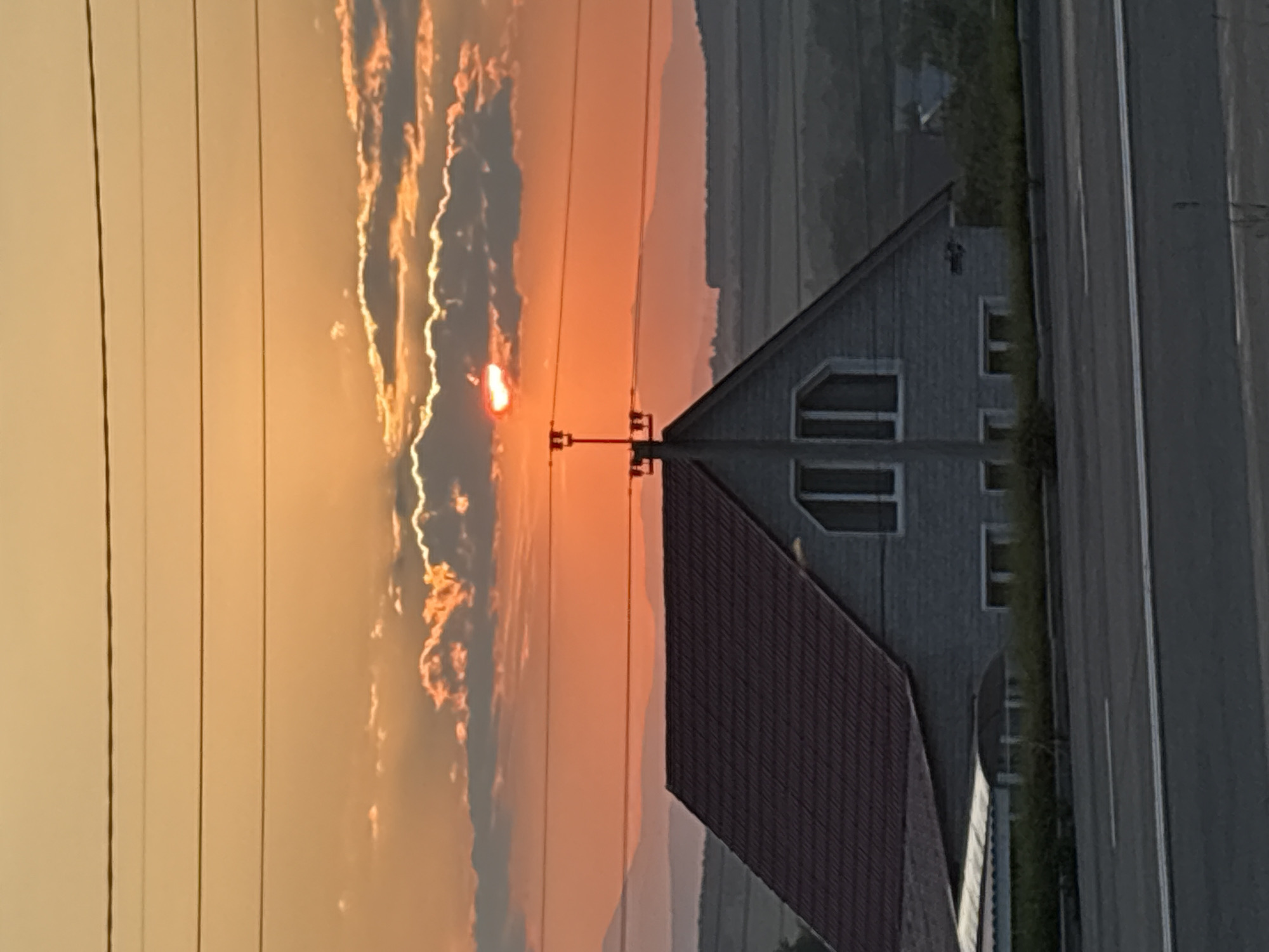 The image depicts a house with a red roof situated in front of a landscape under a sunset sky. The sky is a vibrant orange with clouds tinged with red and yellow. The sun is partially obscured by clouds, casting rays of light. In the foreground, a road is visible. Several power lines stretch horizontally across the upper portion of the image. The house appears to be two stories tall and is constructed of light-colored siding. There are multiple windows on the house, including two distinct multi-paned windows on the upper level. A dark-colored shed-like structure is attached to the left side of the house. The landscape behind the house consists of rolling hills and distant mountains, appearing hazy in the sunset light. The scene appears to be in a rural or semi-rural area, given the presence of open land and the style of the house.