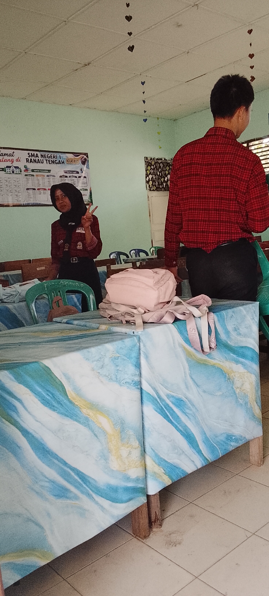 In a classroom setting, two individuals are visible. A young woman wearing a black hijab and a red checkered shirt is standing in the foreground, holding her hand up in a peace sign. She is looking towards the camera. Behind her, to the right, a person wearing a red checkered shirt and dark pants stands with their back to the camera, facing away from the viewer. The room appears to be decorated with hanging paper hearts, and a bulletin board displaying school information is on the wall to the left. Tables covered with a blue and white marbled fabric are in the foreground, with a pink backpack on one of the tables. The floor appears to be tiled. While the specific location within Tanjung Setia, Indonesia, is not visually identifiable from the image alone, the context suggests it is a school.