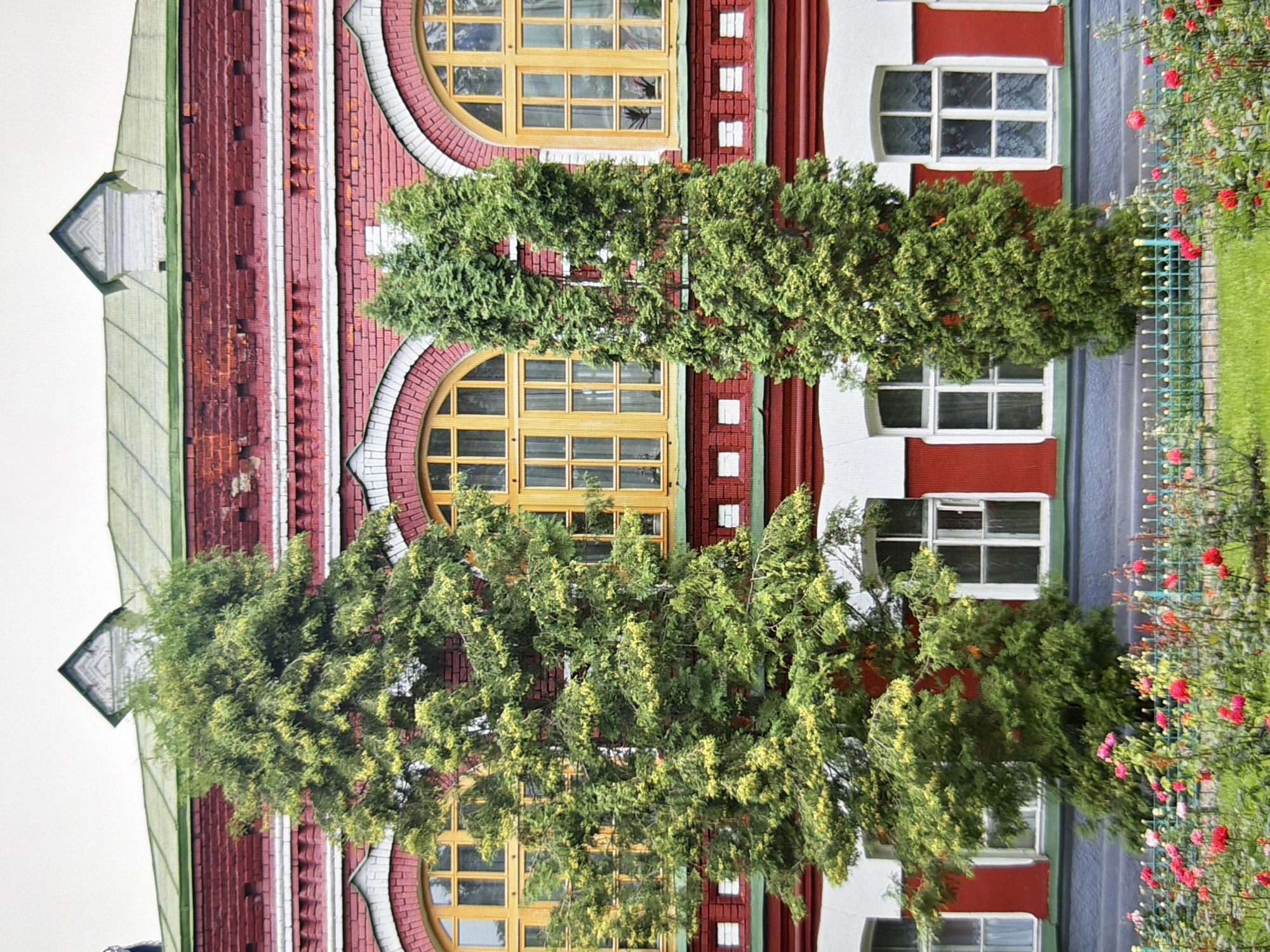 The image shows a section of a red brick building with arched windows and a green roof. Two tall, conical evergreen trees stand in front of the building, partially obscuring the windows. In the foreground, a garden with blooming red and pink flowers is visible, separated from the building by a green metal fence. The scene appears to be a well-maintained garden in front of an old building, possibly part of a historical site or institution. The overall impression is one of tranquility and natural beauty set against an architectural backdrop. Based on its distinctive architecture, the building is identifiable as the former St. Catherine's Monastery in Kyiv, Ukraine, currently housing the National Pedagogical University named after M.P. Drahomanov.