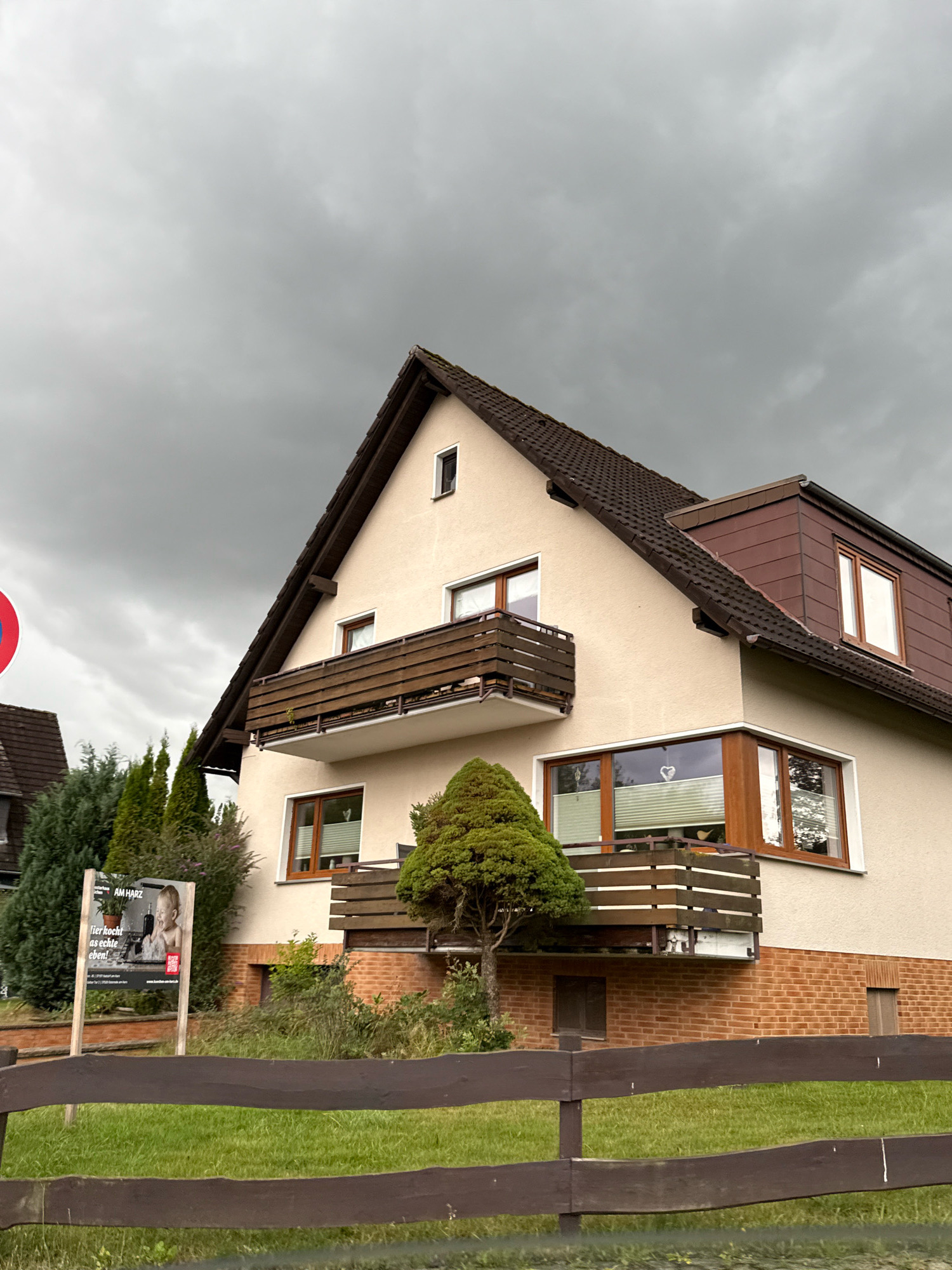 A German house in Bad Sachsa, Germany, is shown under a cloudy sky. The house is light-colored with a dark brown roof and balconies. There are several windows, some with wooden frames. A wooden fence runs across the front of the property, in front of a grassy area. To the left of the house, a sign with a picture of a baby and text in German is displayed. Trees and bushes surround the house.