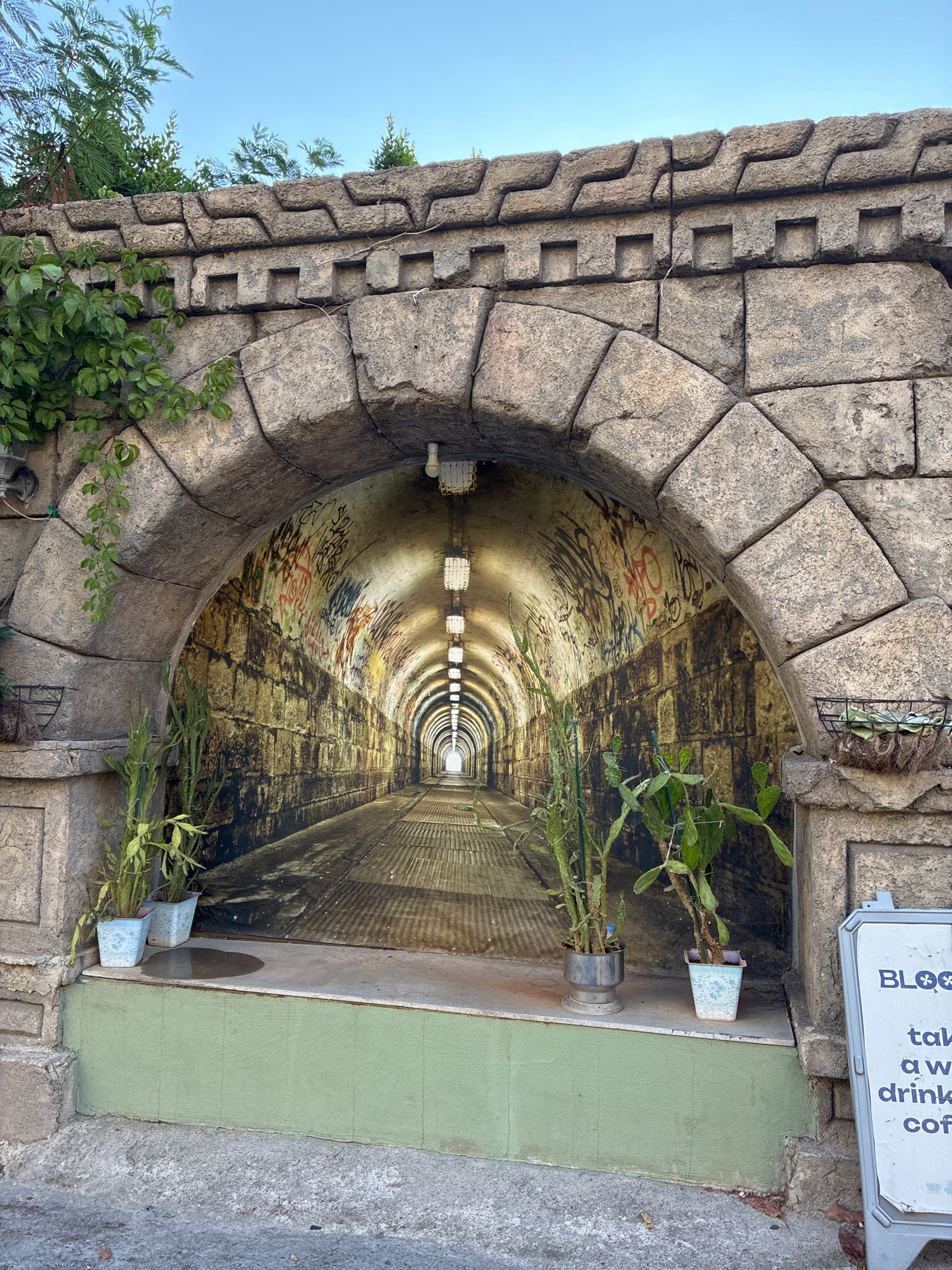 The image depicts the entrance to a tunnel-like structure with a mural on the interior walls, located in Alanya, Turkey. The exterior facade is constructed of rough-hewn stone blocks forming an arched opening. Above the arch, decorative stonework with a zig-zag pattern is visible, interspersed with square indentations. Green foliage drapes down from the top left of the structure.
Inside the archway, the mural creates the illusion of a long, illuminated tunnel. The mural features brick-like walls covered in graffiti, with a perspective view of the tunnel receding into the distance. A series of artificial lights hang from the ceiling of the tunnel, evenly spaced and casting light onto the path. The floor of the depicted tunnel appears to be a metal grate.
In front of the mural, on a raised step painted light green, are three potted plants. The two plants on the left are in light blue and grey pots, while the plant on the right is in a decorative patterned blue and white pot. The plant on the far left has thin, elongated leaves, and the two plants to its right are cactus-like with broader, flatter leaves. A small puddle of water is visible on the step to the left of the plants.
To the right of the entrance, a white A-frame sign with blue text is positioned. The visible text on the sign includes "BLOOM" at the top, followed by "tak", "a w", "drink", and "cof". The ground in front of the structure is a grey, rough-textured surface, likely pavement or concrete. The sky above is clear and blue. The location is identified as Alanya, Turkey.