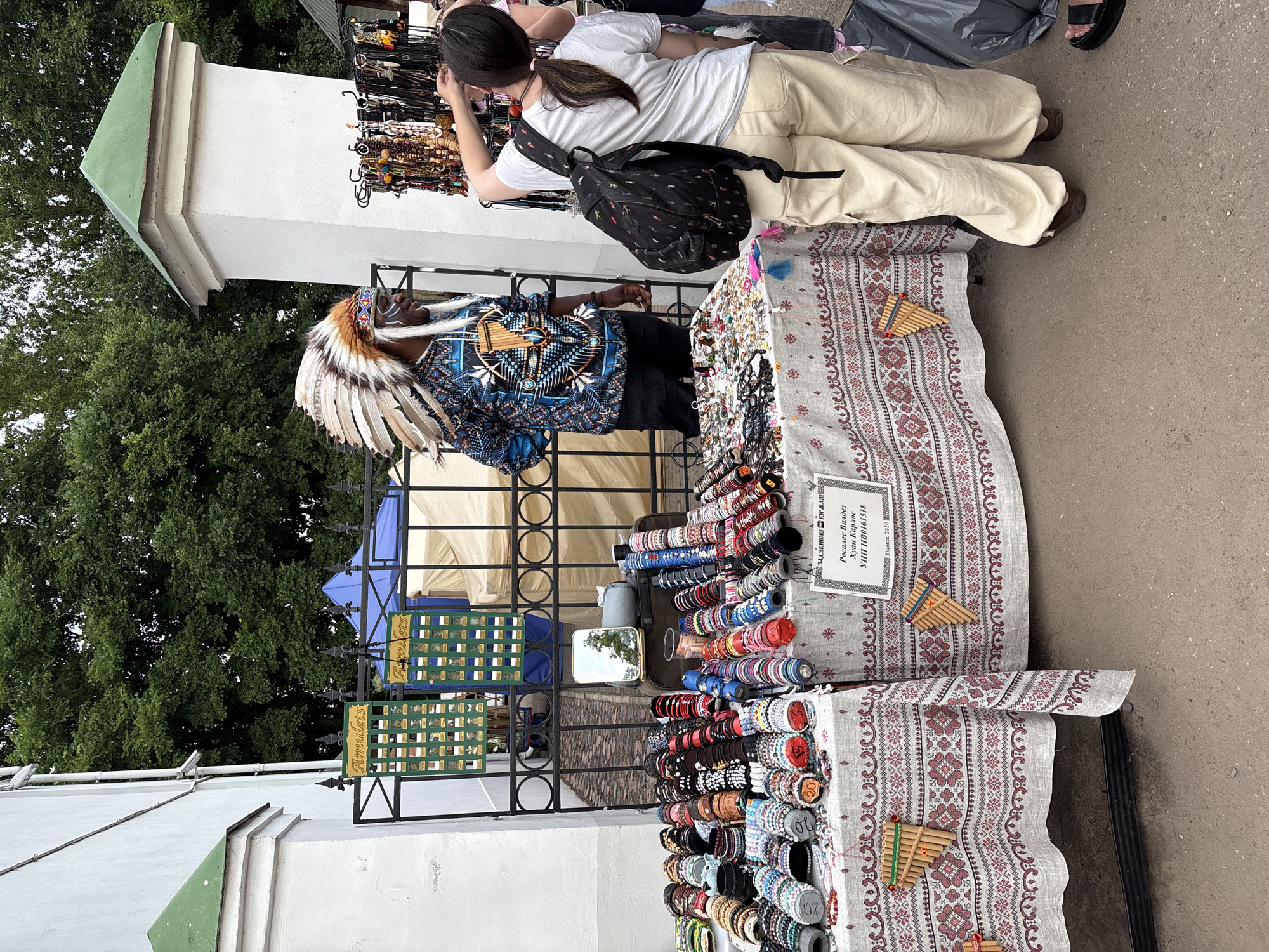 The image depicts an outdoor market scene, likely in the city of Vitsebsk, Belarus, given the Cyrillic text on the sign. A man wearing a Native American-style headdress with feathers and face paint stands behind a table displaying various bracelets and other accessories. The table is covered with a cloth featuring a traditional Slavic pattern. In front of the table, a young woman with a backpack is browsing the merchandise, reaching out to touch an item. Other people are partially visible in the background. The environment includes a white building with a green-tiled roof, a black metal fence, and trees. The ground appears to be a paved surface. A sign with Cyrillic text is visible on one of the tables, indicating potential information about the vendor or products. Another sign with the word "Amulets" is also visible on the fence. The overall scene suggests a cultural or craft market.