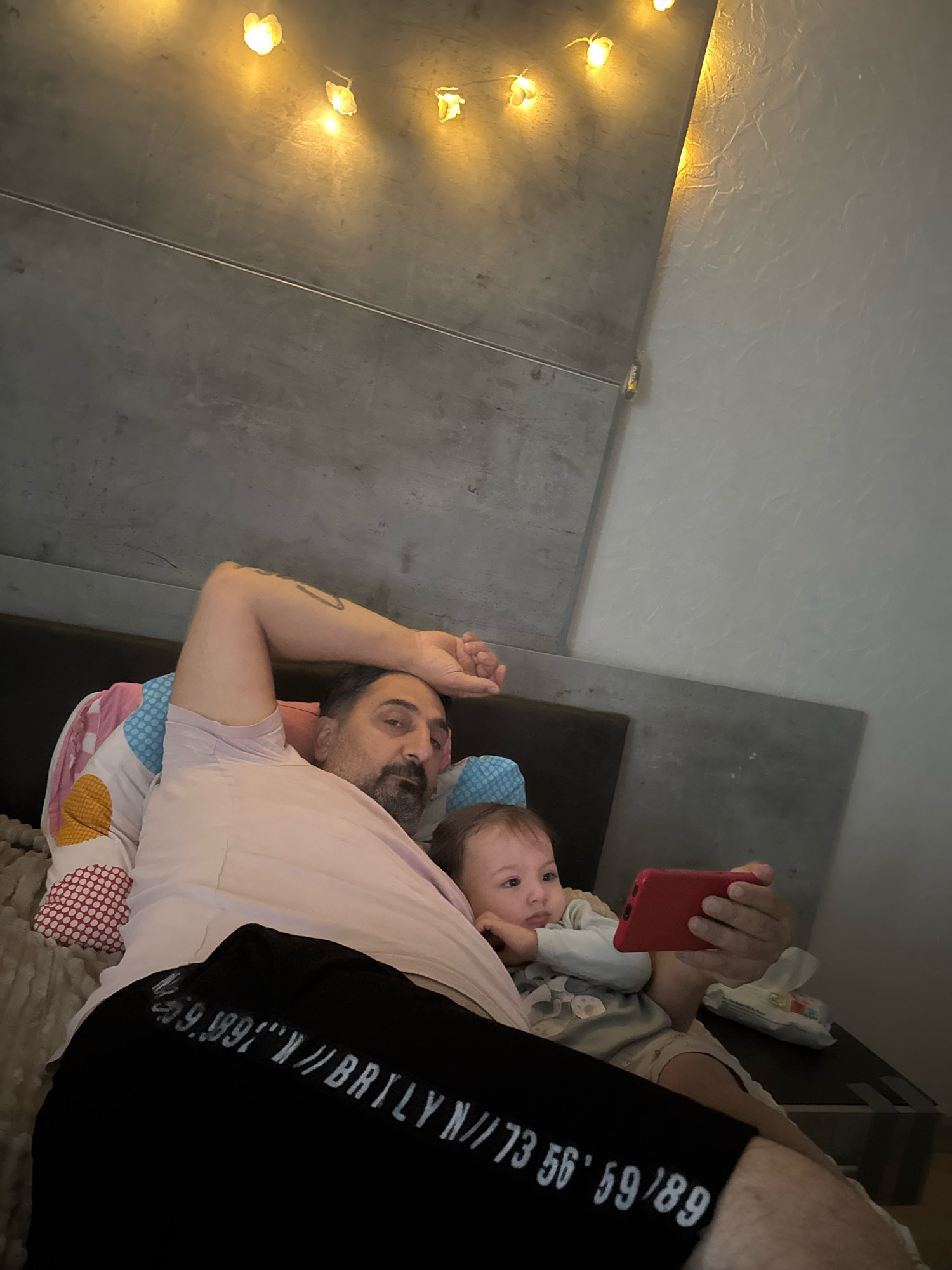A man and a baby are lying on a bed, watching a smartphone. The man is on his side with his arm raised, resting his head on a pillow. He is wearing a light pink t-shirt and black shorts with white lettering. The baby is also lying down, propped up by pillows and watching the phone held by the man. The baby is wearing a light blue onesie. The environment appears to be a bedroom with a grey, textured wall and some string lights overhead. A tissue packet is visible on the edge of the bed. The scene is dimly lit. There is no indication of the location being in Überherrn, Germany.