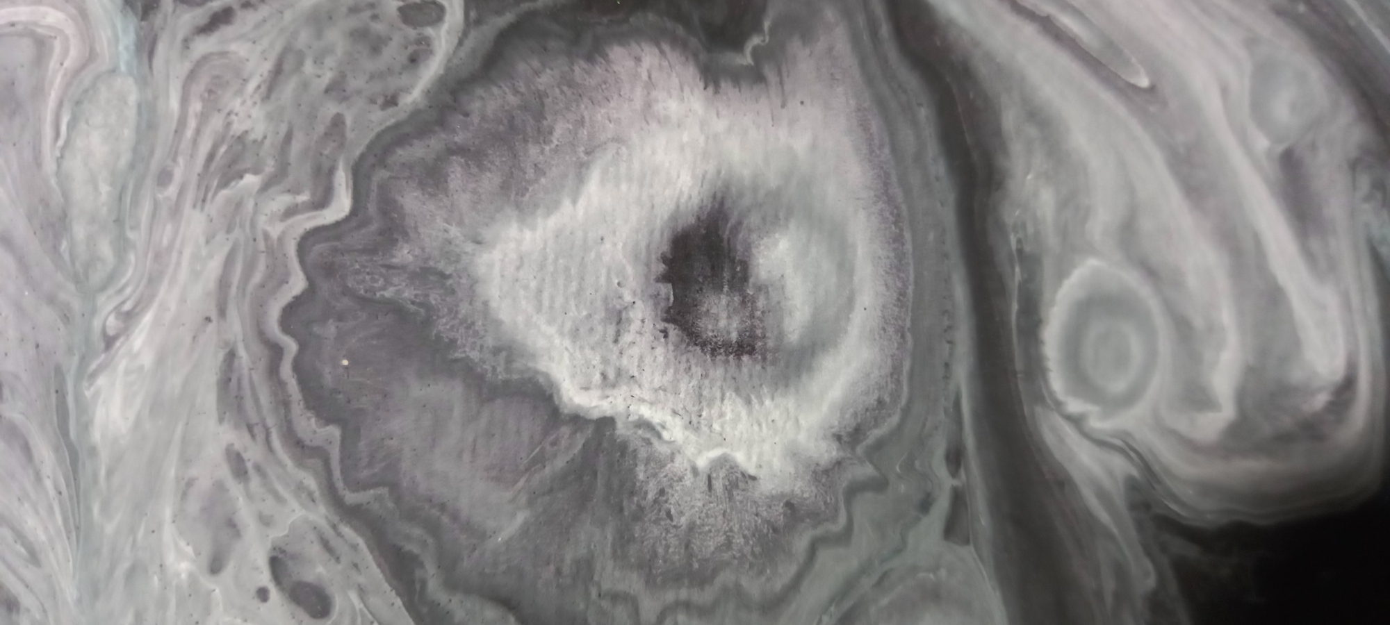 The image depicts an abstract, swirling pattern of black, white, and grey colors, resembling a close-up of a fluid art piece or a natural geological formation like agate. There are no discernible people, objects, or actions visible in the image. The image is a purely visual abstract composition.