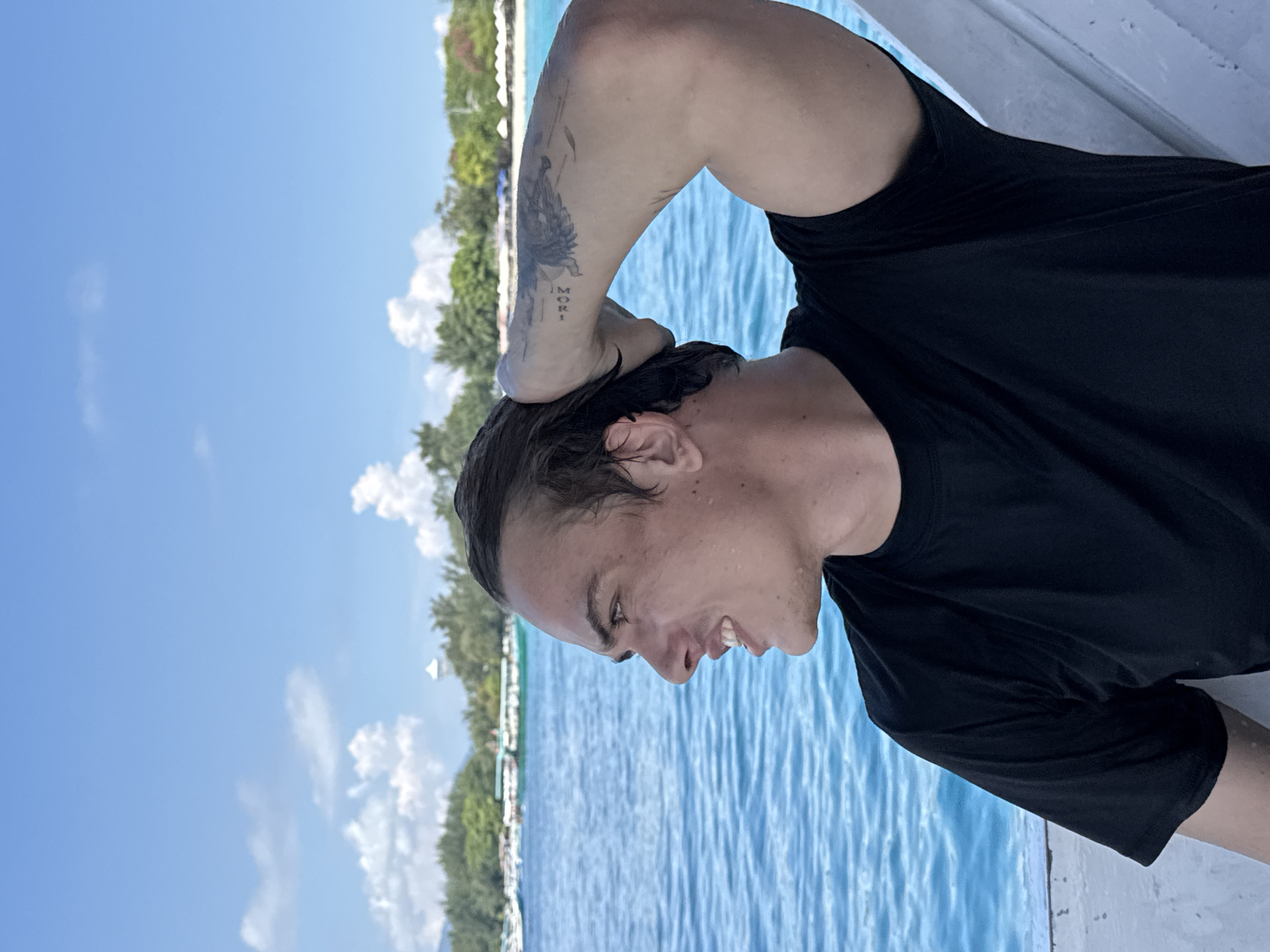 The image shows a man on a boat in Unknown, Indonesia. The man is wearing a black shirt and has a tattoo on his arm. He is smiling and appears to be enjoying himself. The water is turquoise and there are trees in the background.