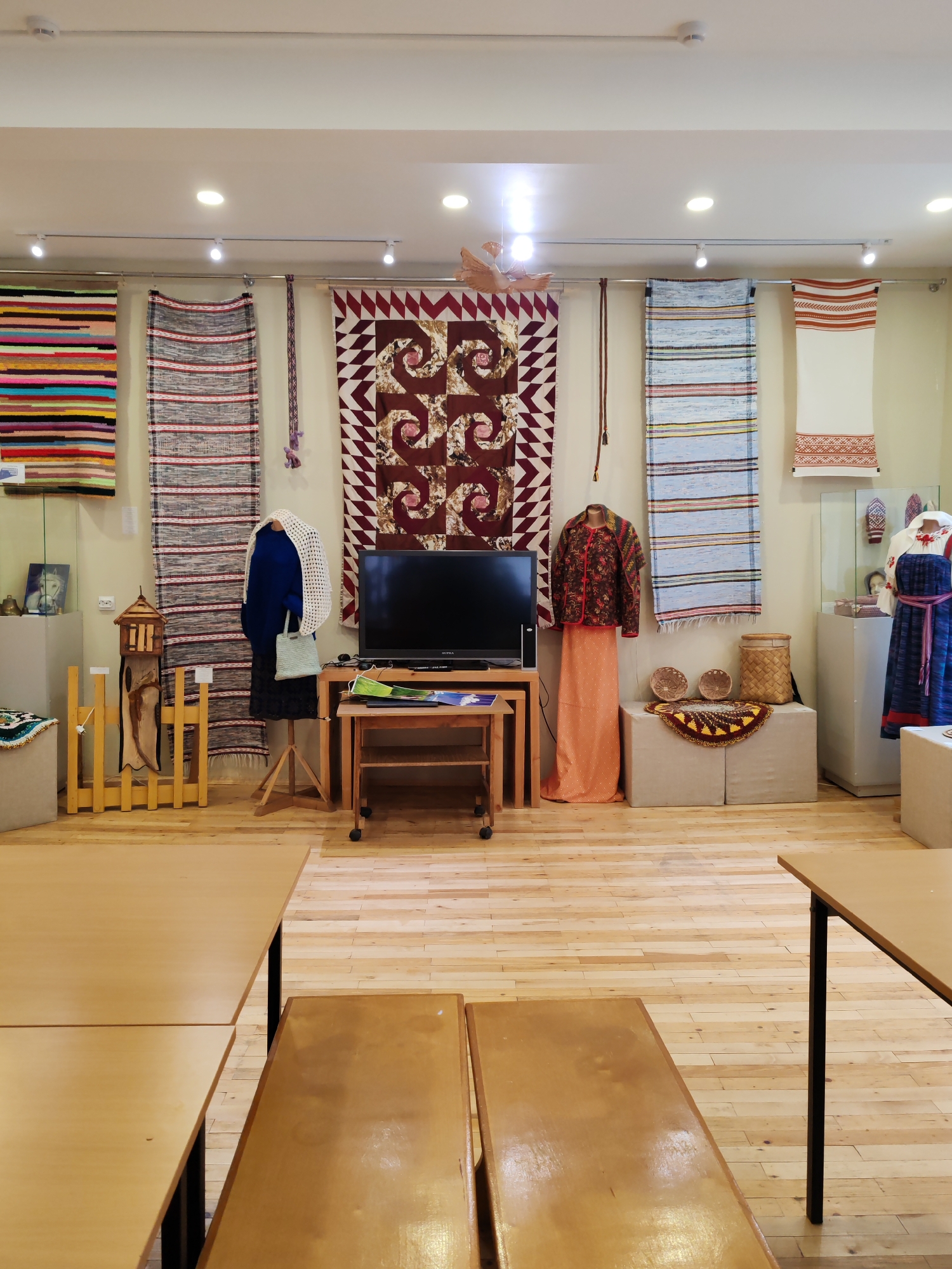 The image depicts an indoor exhibition space, possibly a museum or gallery, with a focus on textile arts. The walls are adorned with various woven items, including colorful striped rugs and intricately patterned wall hangings. In the center of the room, a black television screen sits on a wooden table with wheels, suggesting a multimedia display. To the right, mannequins are dressed in traditional clothing, one in a patterned jacket and an orange skirt, and another in a blue dress with embroidered details. Baskets and decorative textiles are displayed on a raised platform. The floor is made of light-colored wood planks, and the room is illuminated by overhead lighting. In the foreground, two wooden tables are set up, possibly for visitors or workshops. Based on the style of the textiles and the display, this exhibition is likely located in Vizinga, Russia, a region known for its traditional crafts. The scene is static and organized, indicating a curated exhibition. There are no people visible in the image.