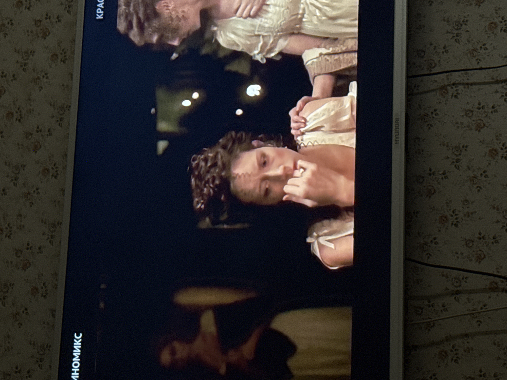 A Hyundai television displays a scene depicting two people, a man and a woman, possibly embracing. The scene is dark, with some lights visible in the background. The woman has curly hair and is wearing a light-colored dress with ribbon detail. The man is positioned above her, also wearing a light-colored garment. The television screen is displaying Russian text at the bottom left corner of the screen which reads "KinoMix". The television itself is placed in an environment with a floral patterned wallpaper, with electrical wires visible behind the TV. The location is unknown and could be Mytishchi, Russia.