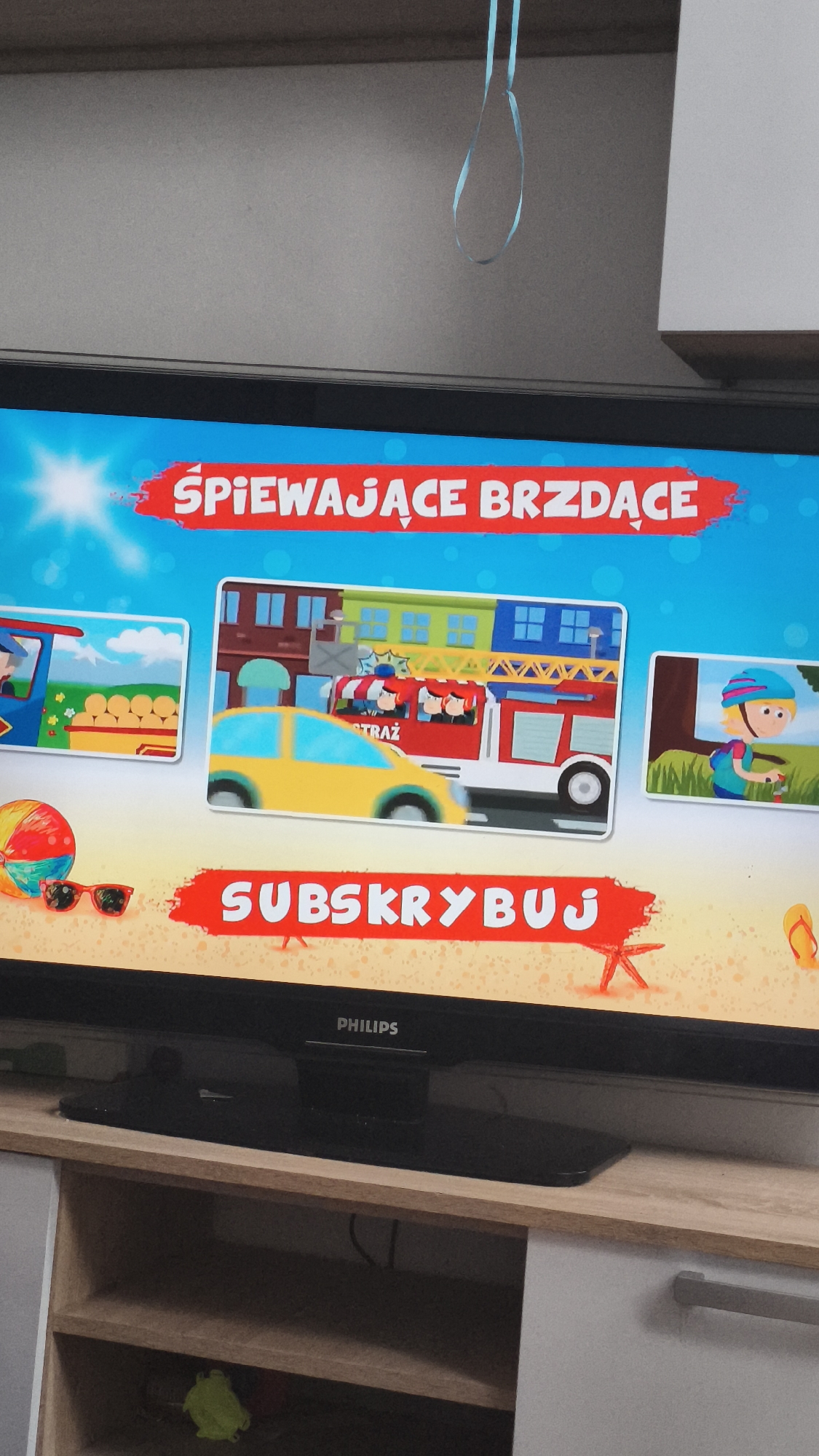 The image shows a Philips television displaying a cartoon animation with Polish text. The title "ŚPIEWAJĄCE BRZDĄCE" (Singing Toddlers) is displayed in red at the top. Below are three cartoon scenes: a train, a fire engine, and a child riding a bike. "SUBSKRYBUJ" (Subscribe) is displayed in red text at the bottom. The television is on a wooden shelf with white drawers, and a blue ribbon hangs down from above the TV. The location of the image is not discernible from the visual information. Thus the location of Nowy Tomyśl, Poland, can neither be confirmed or denied with the data available.