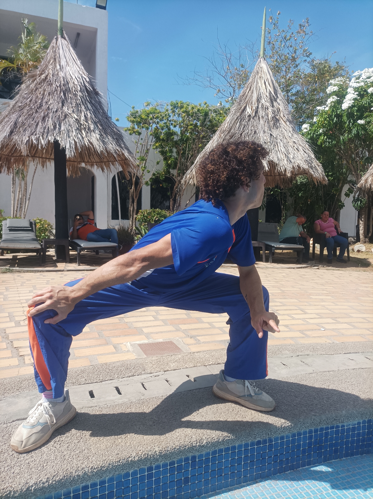 In a sun-drenched outdoor setting in Porlamar, Venezuela, a person with curly brown hair, wearing a royal blue short-sleeved top and matching track pants with orange side stripes, is captured in a dynamic pose. They are performing a wide squat or stretch, with their left hand resting on their knee and their right hand pointing forward, while looking intently towards the right side of the frame. Their light grey and white sneakers are visible at the edge of a swimming pool, which features blue mosaic tiles.
The immediate foreground shows a light-colored tiled patio. In the background, several thatched umbrellas, known as churuatas, cast shadows over lounge areas. Under one churuata to the left, a man in an orange shirt relaxes on a lounge chair, seemingly engaged with a phone. Further right, another man in a light green shirt and a woman in a pink top are seated in chairs, enjoying the relaxed atmosphere.
The environment suggests a resort or hotel, with white buildings, lush green trees, and flowering white bushes contributing to a tropical ambiance. The clear blue sky and strong shadows indicate a bright, sunny day, likely around midday. No explicit text is visible on any subject or object in the image.