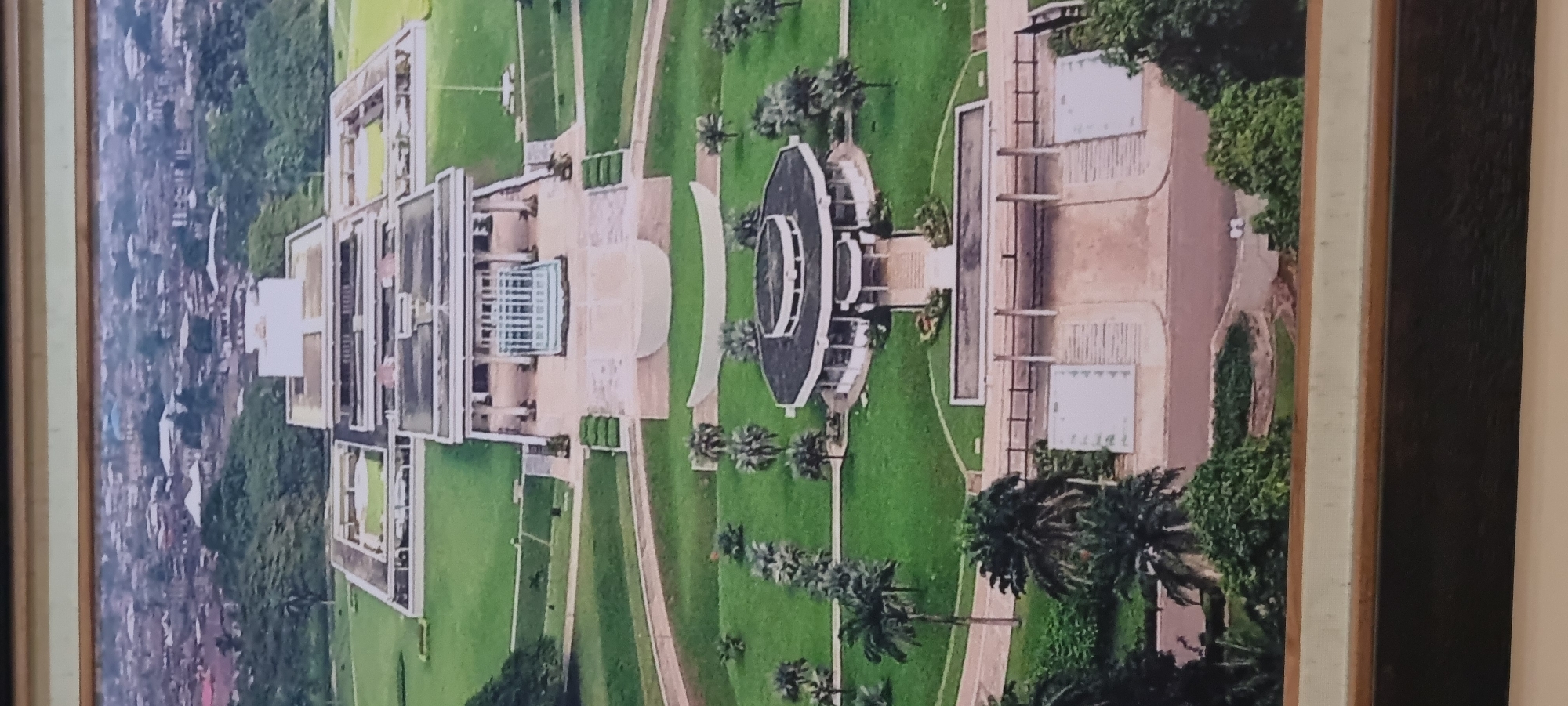 This is an aerial, slightly angled photograph of the sprawling Presidential Villa complex in Abuja, Nigeria. The image captures the vast, manicured grounds surrounding the main presidential residence, which is a modern, rectangular building with multiple levels and flat roofs. Green lawns dominate the landscape, punctuated by pathways, trees, and decorative elements. In the foreground, a circular building with a dark roof and white walls is visible. Further back, the main presidential structure is seen with a large, open plaza in front of it, featuring a central water feature or fountain. Beyond the complex, the dense urban sprawl of Abuja is visible, with numerous buildings and the natural topography of the area. The overall impression is one of grandeur and scale, showcasing the administrative heart of the Nigerian government.