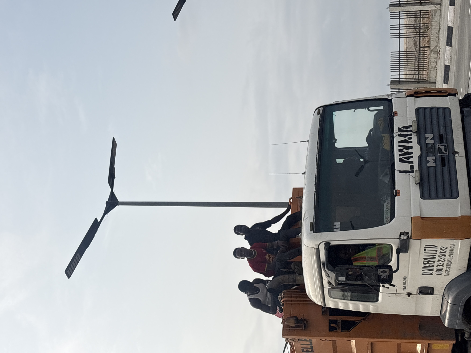 In Alaguntan, Nigeria, three young Black men are seated on the top rear of an orange garbage truck. The man on the left wears a white tank top and dark pants, the man in the middle wears a red shirt and dark pants, and the man on the right wears a black shirt and dark pants. The truck has "D. NIGERIA LTD" printed on its side with a phone number and email address. The truck also has "LAWMA" printed on the front. The truck is on a road with a fence in the background. There are tall streetlights in the background. The sky is overcast.