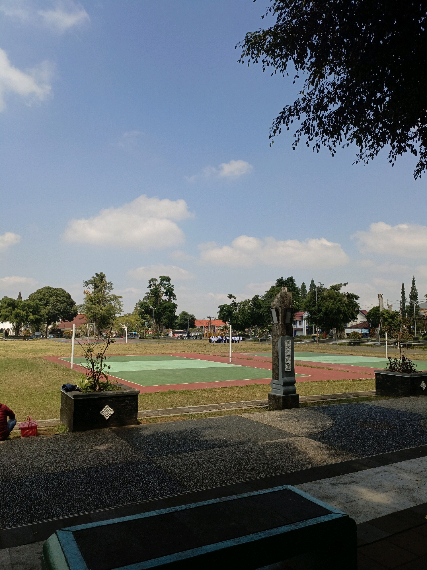 In an outdoor, open-air scene in Wonosobo, Indonesia, on a clear day with scattered white clouds, the foreground features a dark grey, textured paved area with a light blue-edged bench in the lower right corner. Adjacent to this paved area are two dark, rectangular planters. The planter on the left contains a small, green plant.
Beyond the planters is a grassy field with two distinct sports courts. The court closer to the viewer is divided into two sections, one green and one red, bordered by a red perimeter. A white pole stands on the left side of this court. Further back, another similar court is visible, also divided into green and red sections.
In the middle ground, a stone monument, dark grey and featuring intricate carvings, stands between the two sports courts. A line of trees with lush green foliage forms a backdrop behind the sports fields. To the left of the scene, a white building with a dark, steepled roof is partially visible among the trees. In the far distance, several other buildings and more trees are scattered across the landscape. A group of people, some wearing white uniforms, are standing near the far sports court, appearing to be in a line or formation. A person in a red jacket is seated on the ground to the left of the planter on the far left, with a red basket beside them. The scene conveys a sense of a school or recreational facility in a semi-urban environment.