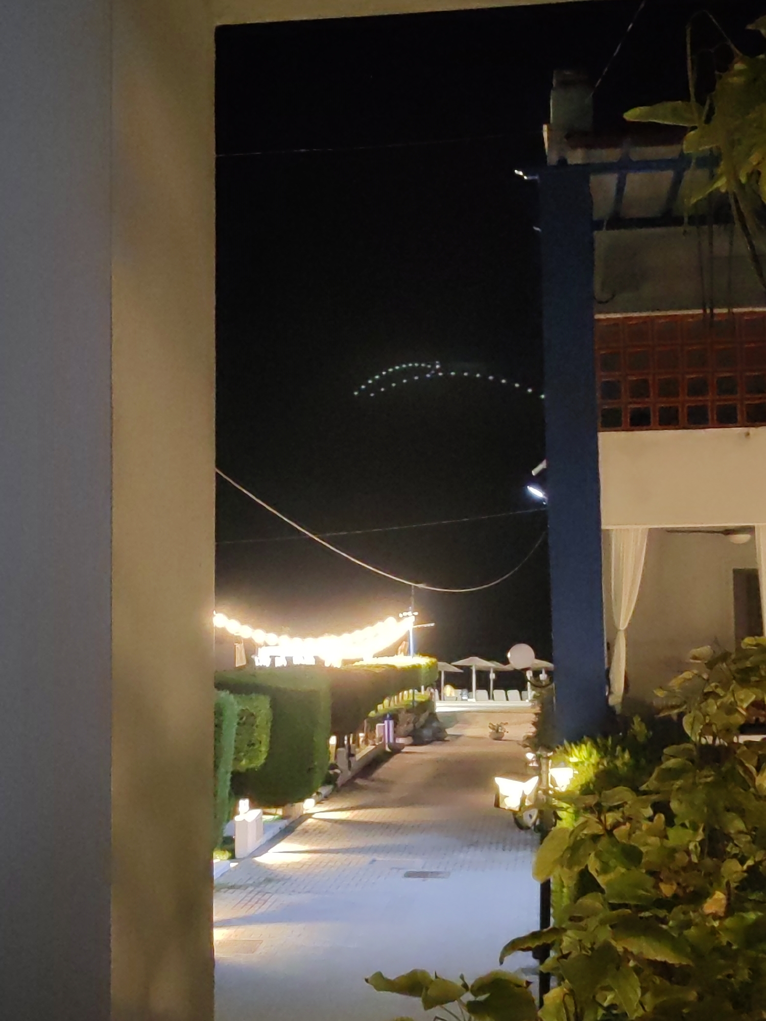 An outdoor nighttime scene is captured from behind a cream-colored column on the left, looking toward a resort-like area. The sky is dark, with a series of white lights forming a curved pattern high up, possibly an airplane's contrail or a string of lights. Below this, a string of warm-toned lights arcs across the scene at a lower elevation, illuminating the pathway. The pathway is paved with light-colored stones and lined with neatly trimmed green hedges. Several white umbrellas and lounge chairs are visible in the background, suggesting a pool or seating area. A building with blue and orange architectural elements is on the right side of the image, with white curtains visible in an open doorway. Lush green foliage is in the foreground on the right, partially obscuring the view. The lighting is artificial, primarily from the string lights and spotlights illuminating the hedges and pathway.