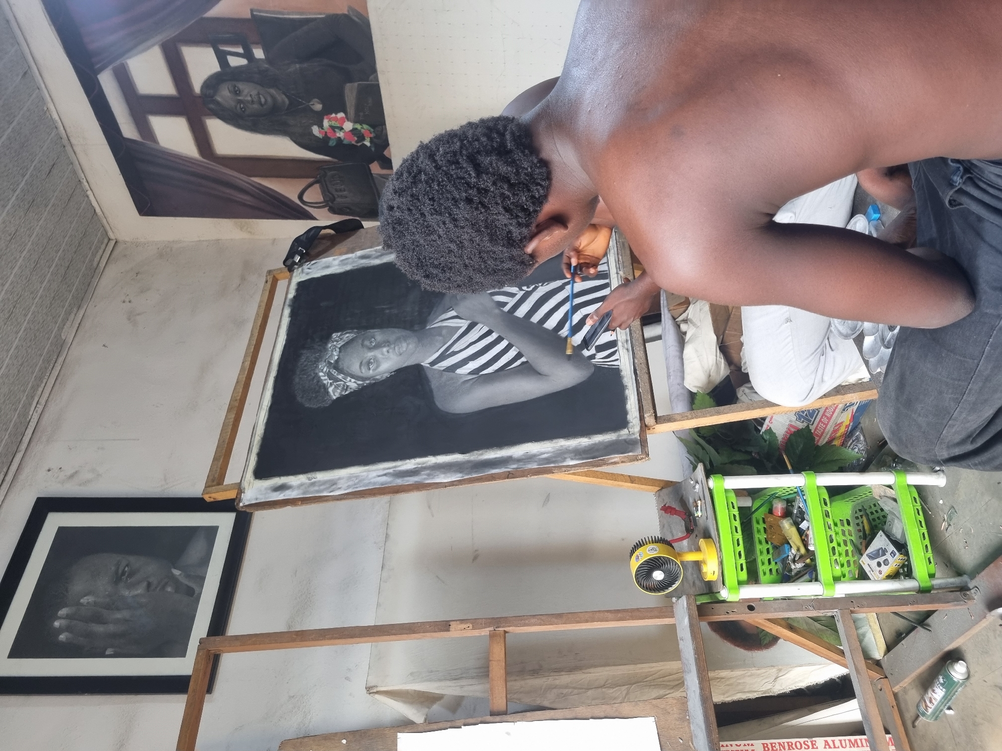 In Onitsha, Nigeria, a bare-chested artist with dark hair, bent over, meticulously works on a black and white portrait with a blue brush. The portrait features a woman in a striped shirt. The artist is surrounded by his workspace: other finished artwork hangs on the walls, including a larger piece featuring a woman with flowers, and a framed portrait. A rickety wooden structure supports the artist's work and tools. Cluttered within the structure are green baskets filled with art supplies, and a small yellow fan. Other items, including leaves, an aerosol can, and packaging, are scattered nearby.