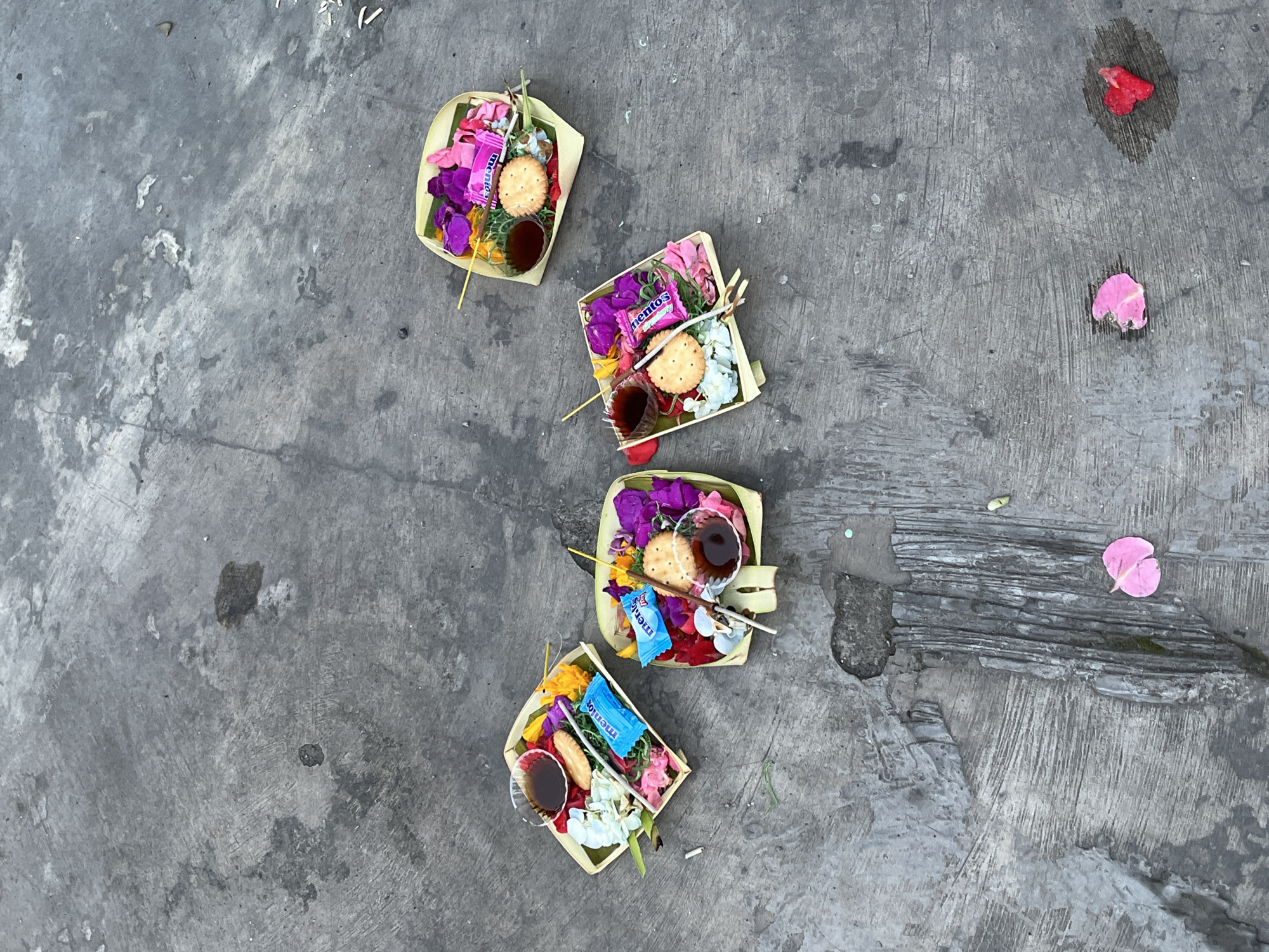 The image depicts a section of concrete ground in Baturiti, Indonesia, featuring four small, square woven trays. Each tray contains a collection of items including flowers (various colors including purple, pink, white, and yellow), a biscuit, a small cup of dark liquid, and a package of Mentos. Scattered nearby are a few fallen flower petals. The ground is cracked and stained.