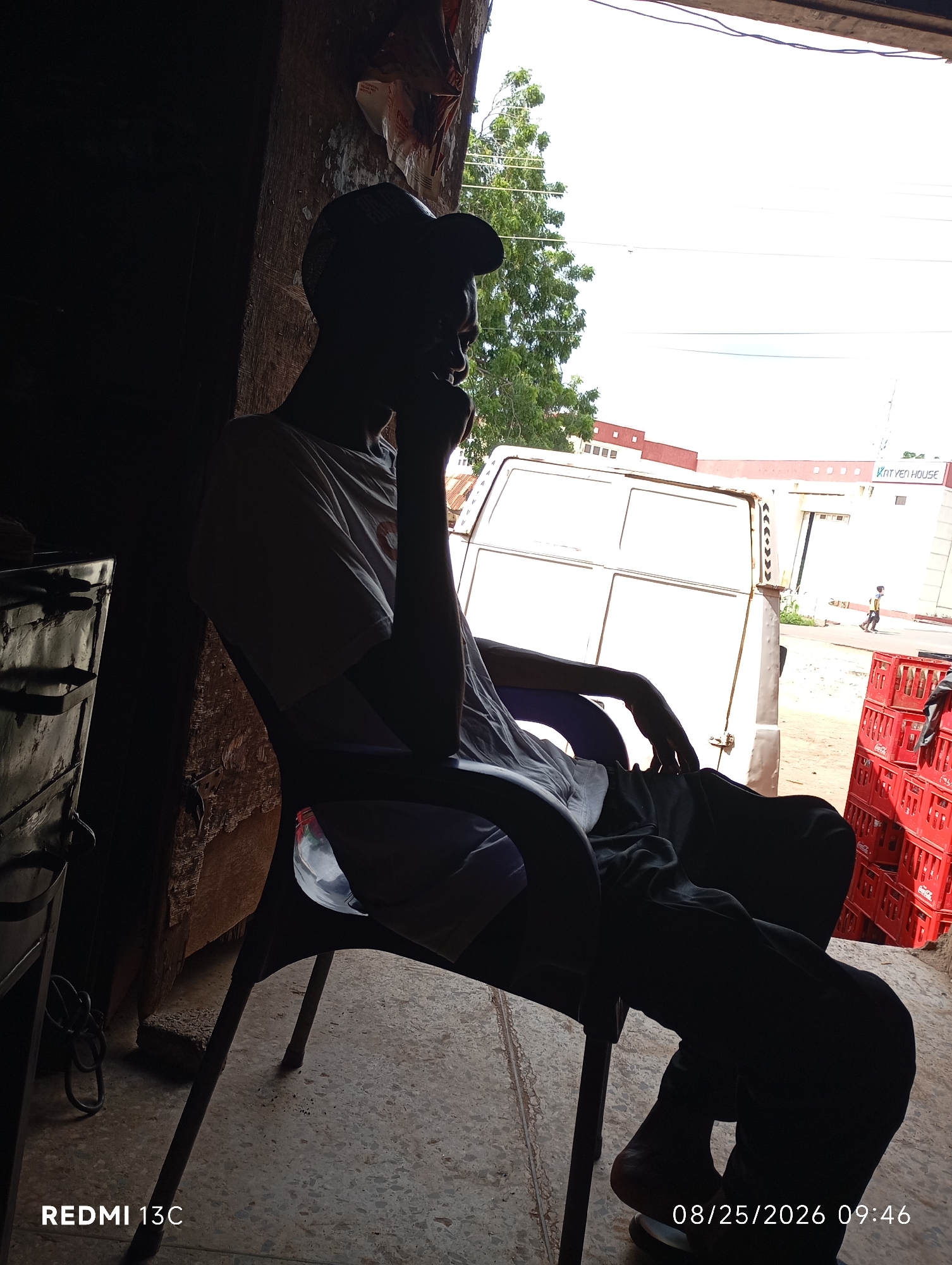 The image shows a silhouetted young man wearing a baseball cap and a t-shirt, sitting in a chair in what appears to be an outdoor shaded area. He is looking to the right, with his hand near his face, suggesting he may be in thought or observing something. Behind him, a white van is visible, along with a building in the background that has "KAT YEN HOUSE" written on it. To the right of the van, there are stacked red crates, possibly containing beverages. The scene suggests a commercial or roadside setting. The foreground shows a dark, textured surface, possibly a floor or ground. The image also includes a timestamp "08/25/2026 09:46" and "REDMI 13C" in the bottom left and right corners respectively. The location is identified as Shendam, Nigeria.
