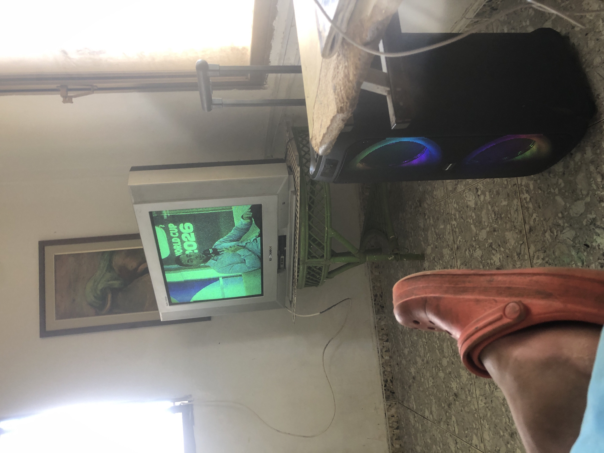 The image depicts an indoor scene, likely a living room, with the camera angle focused on a person's foot wearing an orange Croc shoe, positioned in the foreground. To the left, a window with a sheer curtain lets in bright natural light, partially obscuring the wall. In the midground, an older model television set displays "WORLD CUP 2026" with a person speaking into a microphone. The television rests on a green metal table with intricate scrolling details. Behind the television, a framed picture hangs on the wall, showing an image that appears to be an elephant. To the right, a large black speaker with colorful LED lights is visible, and a portion of a wooden table with a rough surface. The floor is tiled with a gray and beige pattern. The overall environment suggests a casual, lived-in space. There is no information in the image to indicate the location is specifically in La Tagua, Venezuela.