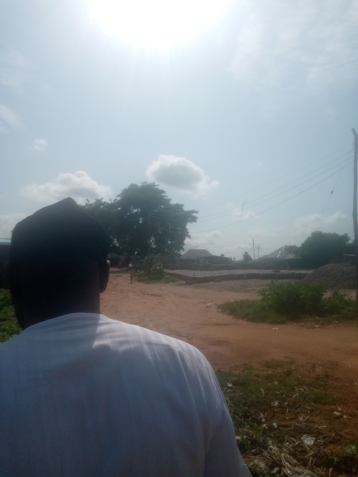 The image depicts an outdoor scene on a bright, sunny day in Jalingo, Nigeria. The perspective is from behind a person wearing a white shirt and a dark cap, facing away from the camera. The background shows a reddish-brown dirt road with patches of dry grass and some scattered debris. In the mid-ground, there are stacks of what appear to be bricks or building materials, and several low-rise structures, possibly homes or workshops, with corrugated iron roofs. Lush green trees are visible, one large tree dominating the left side of the frame. Overhead, power lines stretch across the sky, supported by a wooden pole on the far right. The sky is light blue with scattered white clouds, and the sun is prominently visible, causing a bright glare in the upper portion of the image. The overall environment suggests a rural or semi-urban setting within Jalingo.