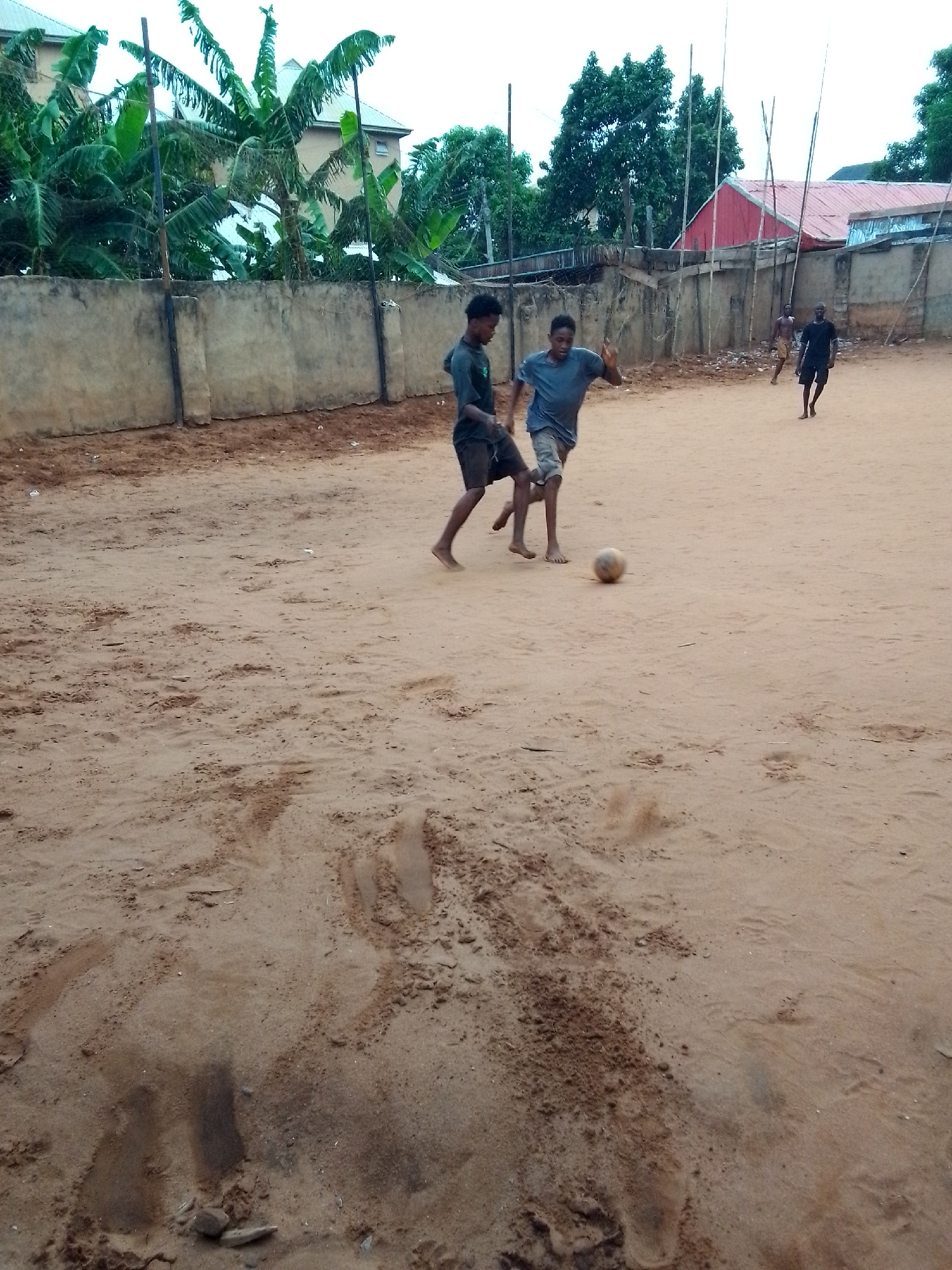 The image depicts an outdoor scene of four young males playing soccer on an unpaved, sandy dirt field. In the foreground, two players are central to the action; one wears a dark green or black shirt and shorts, and the other wears a grey shirt and shorts. Both appear to be barefoot or have minimal footwear. A light-colored soccer ball is positioned near their feet, indicating active play. In the mid-ground and background, two additional male figures are visible, observing or participating in the game; one wears a dark shirt and shorts, and the other is partially obscured.
The setting is an informal playing ground with a rough, uneven sandy surface marked by footprints and disturbed patches of dirt. Along the back of the field, a low, weathered concrete wall separates the playing area from dense green tropical vegetation, likely banana or plantain trees, and several buildings with corrugated iron roofs, one of which is distinctly red. Scattered around the edges of the field are thin, tall poles, possibly made of wood or bamboo, which may serve as makeshift goalposts or boundary markers.
The time of day appears to be daytime, likely late afternoon, under an overcast or hazy sky that casts soft, diffuse light across the scene, resulting in minimal distinct shadows. The informal nature of the game and the environment suggest a casual, community-level sporting activity. There is no visible text in the image. The scene conveys a sense of everyday life and recreation in an informal, semi-urban or rural setting.