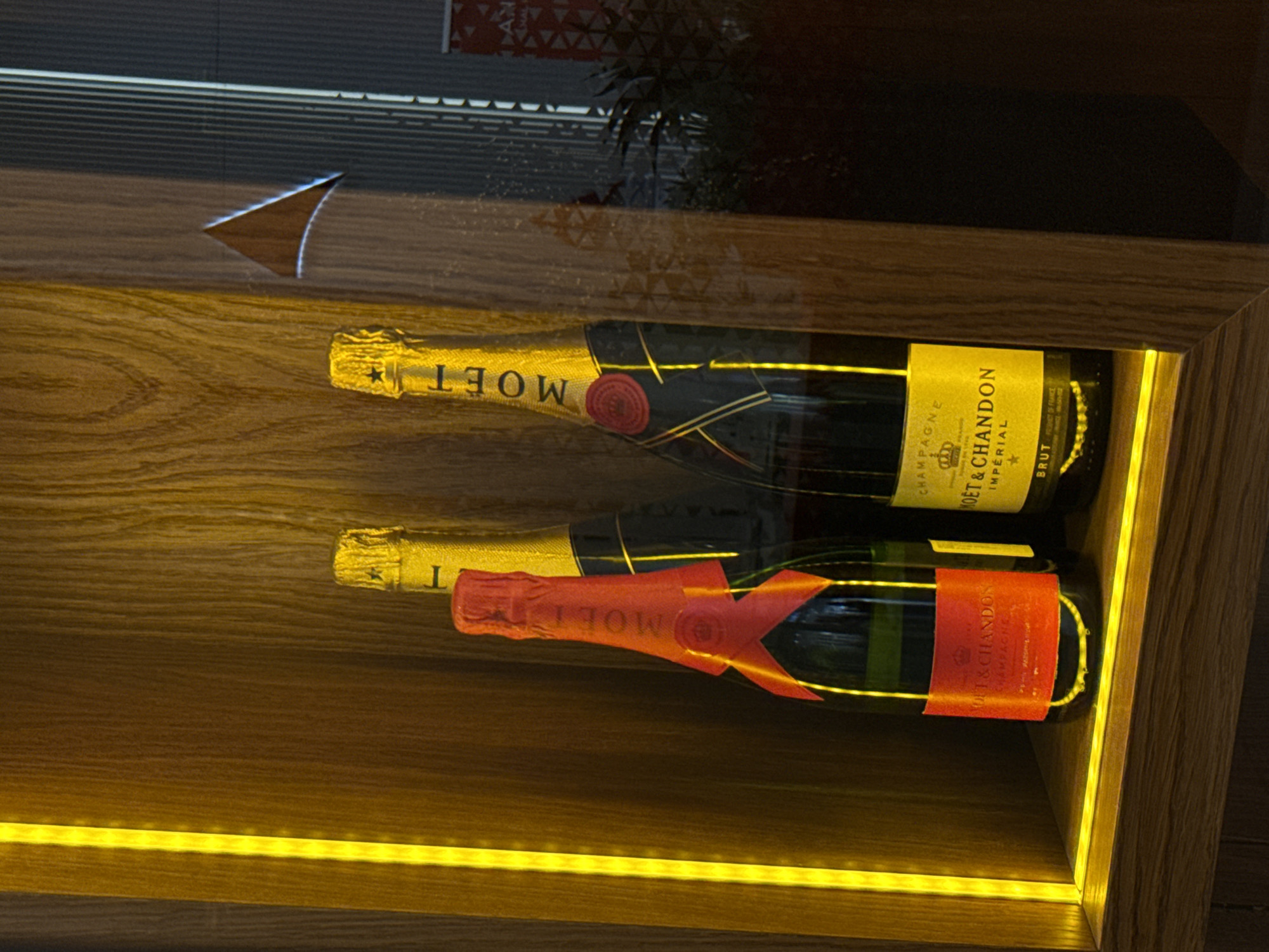 Two bottles of Moët & Chandon champagne rest on a wooden shelf illuminated by a yellow LED strip. The top bottle is the classic Imperial Brut, while the bottom one is the Rosé version. The shelf appears to be within a dimly lit space, possibly in a retail establishment or bar in Belgrade, Serbia. The reflection on the shelf's surface suggests the presence of an additional structural element, possibly a window or another shelf, behind it.
