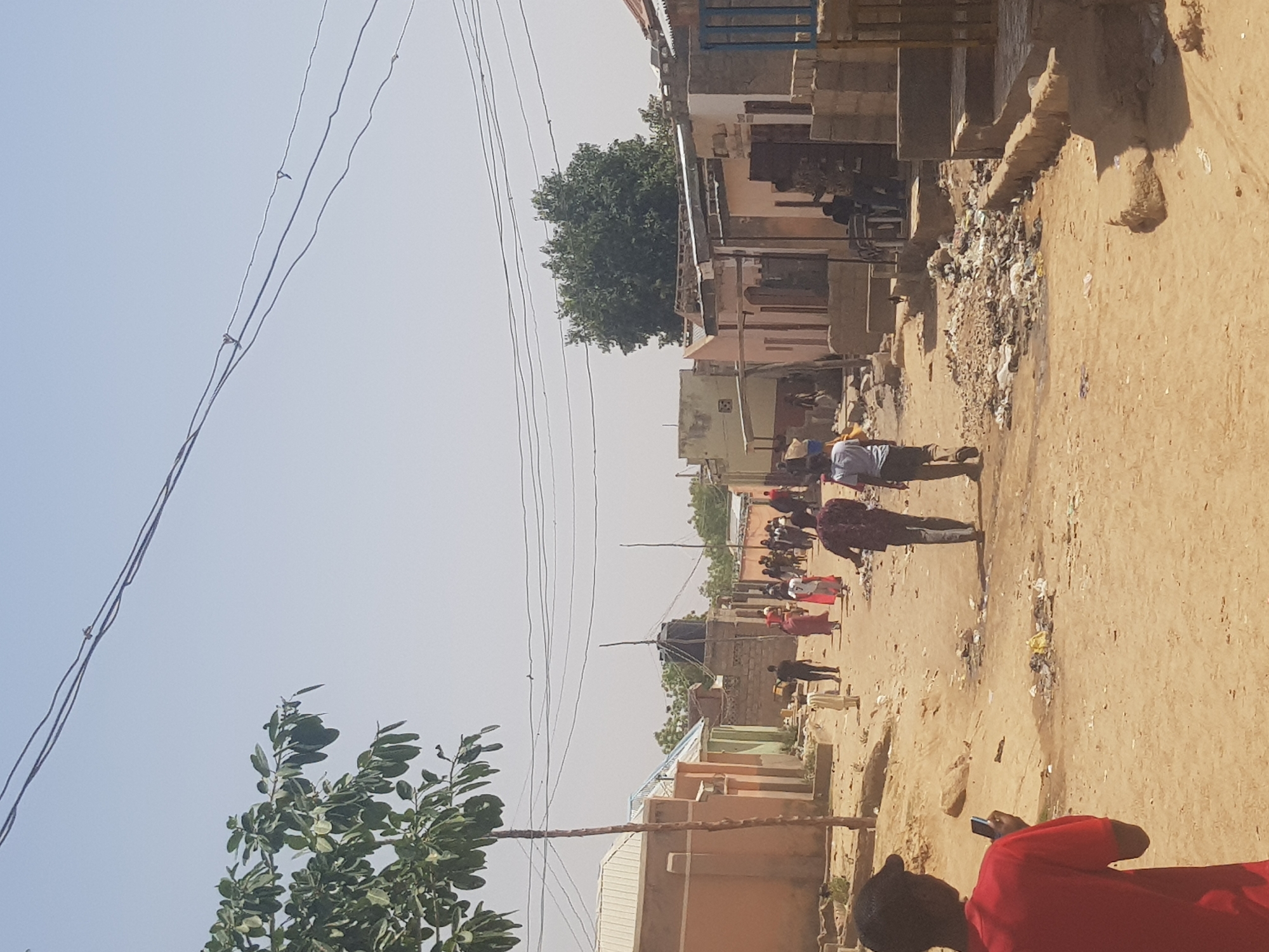 The image shows a street scene in Dabawa, Nigeria. The street is unpaved and sandy, with buildings lining both sides. Debris and litter are scattered along the street's edge. Several people are walking down the street. Power lines are visible above. One individual is in the foreground, partially visible, wearing a red shirt and holding a mobile phone.