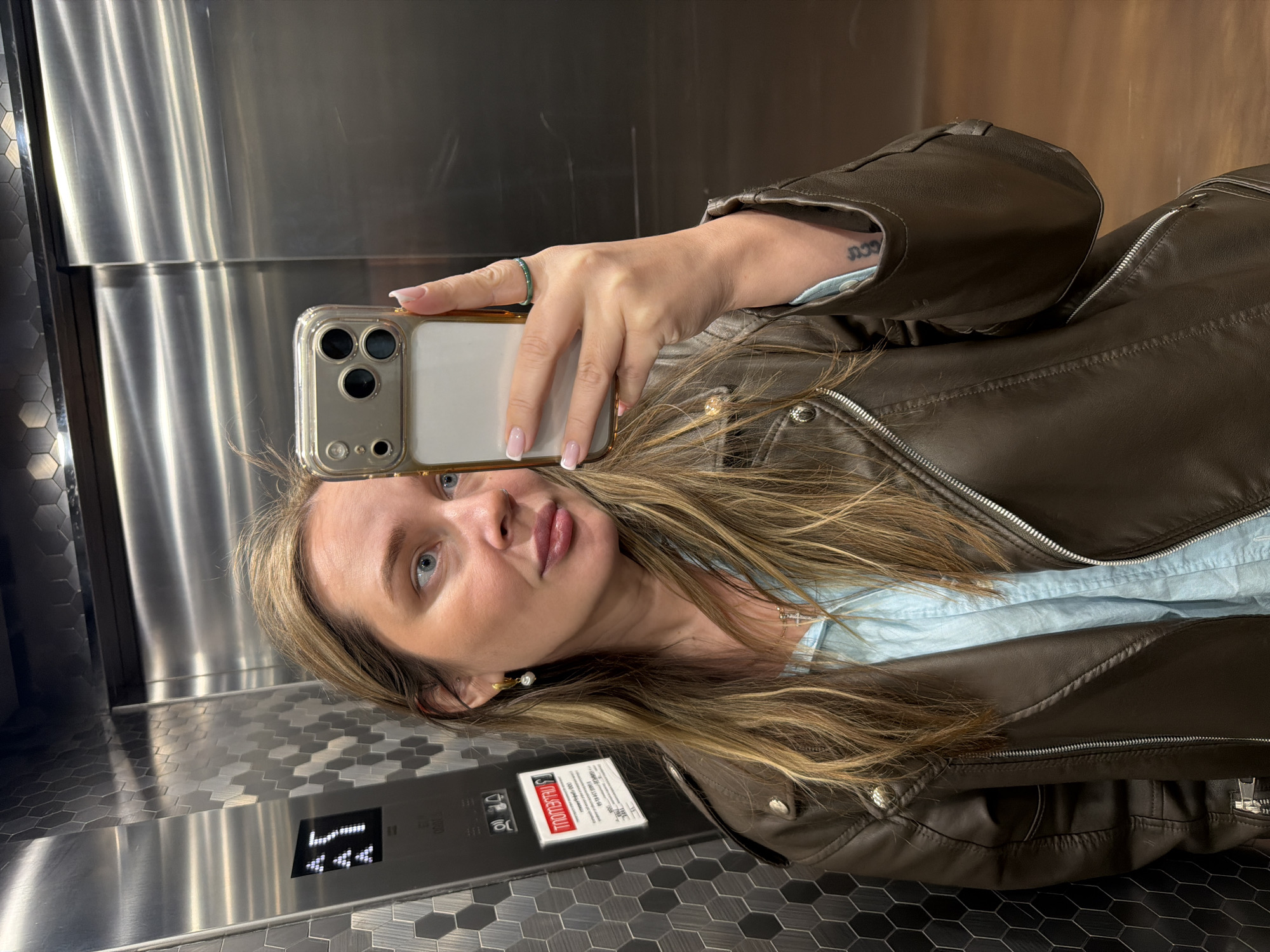 A woman is taking a selfie in an elevator. She is wearing a brown leather jacket over a light blue shirt, a ring, and earrings. The elevator's control panel is visible, showing that the elevator is on the 7th floor. The elevator interior consists of stainless steel and dark grey honeycomb paneling. The location is not identifiable as Moscow, Russia.