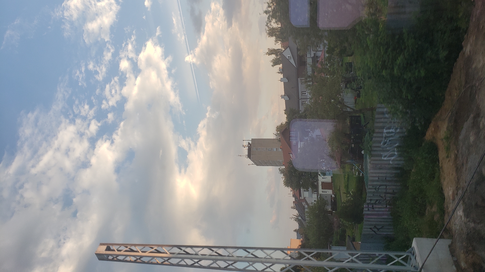 The image depicts a view of the city of Poprad, Slovakia, during what appears to be late afternoon or early evening, given the lighting conditions. In the foreground, a tall, metallic lattice structure, possibly a pylon or part of a communication tower, is visible on the left side of the frame. It is made of crisscrossing metal beams and stands on a concrete base.
Beyond the lattice structure, a residential area is visible. Several buildings are present, including a prominent, multi-story apartment building with numerous windows and what appears to be antenna equipment on its roof. This building is situated behind a row of smaller houses with pitched roofs. The houses are painted in neutral colors and are interspersed with trees and greenery. A section of a corrugated metal fence with graffiti on it is also visible in the lower part of the image, separating the foreground from the residential area.
The sky is a prominent feature, displaying a mix of blue sky and clouds. Some clouds are white and puffy, while others are more diffused and tinted with the warm hues of sunset. A contrail from an aircraft is visible stretching across the sky. The overall impression is a calm, urban landscape under a dynamic sky. There are no people or animals visible in the scene.