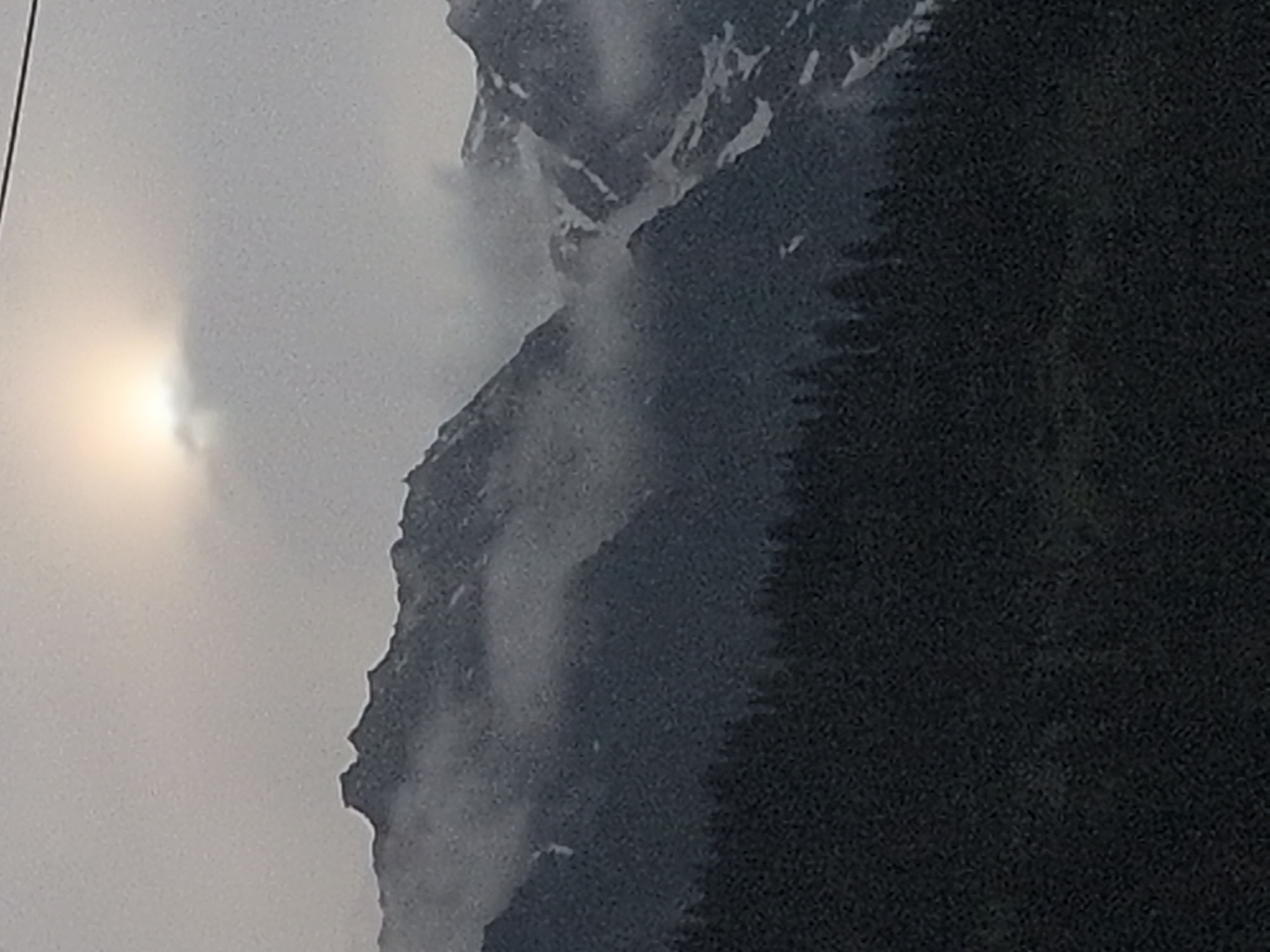 The image shows a hazy, overcast sky with a bright, diffused sun partially obscured by clouds. Below the sky, a dark, dense forest of evergreen trees covers the lower portion of the frame. Rising above the forest are rugged, mist-shrouded mountains. Some of the mountain peaks appear jagged and rocky, while others are covered in patches of snow. The overall atmosphere is moody and somewhat subdued, suggesting a cool or damp climate, possibly at a higher elevation. The scene is likely a natural landscape, given the presence of mountains and a forest. The provided context suggests this view is from Vashisht, India, a place known for its mountainous terrain and natural beauty.