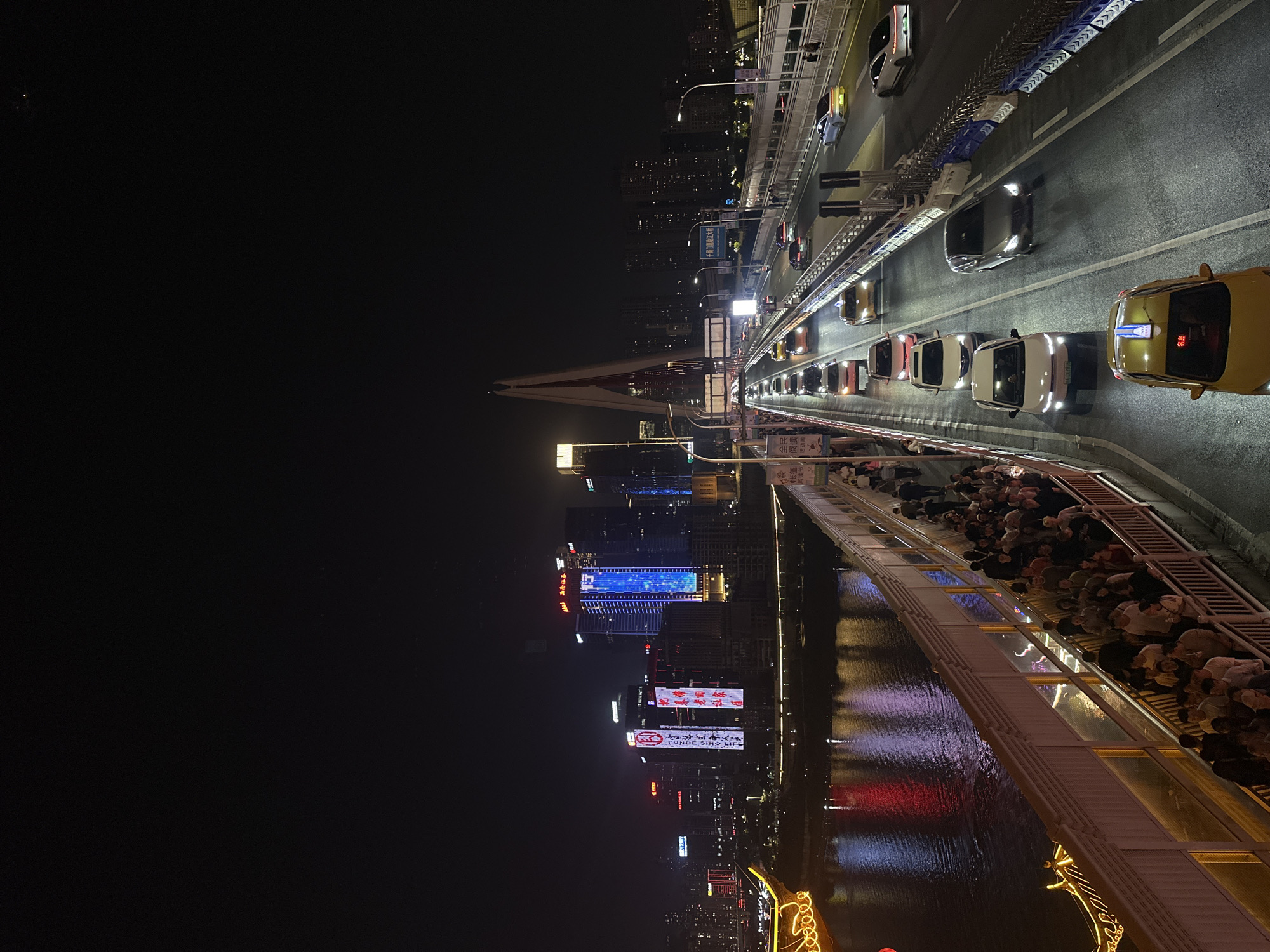 This nighttime image captures a bustling urban scene in Yuzhong District, China, centered around a brightly lit bridge. The perspective looks down onto the bridge and the adjacent cityscape, which stretches into the dark horizon.
The main subject is a multi-lane bridge carrying heavy traffic. Numerous cars, including several yellow taxis and other private vehicles, are visible in a queue, their headlights and taillights illuminating the road surface. A blue and white barrier system runs along the center of the bridge road. Alongside the vehicle lanes, a wide pedestrian walkway is densely packed with people, suggesting a popular viewpoint or a busy thoroughfare.
To the left of the bridge, a modern city skyline emerges from the darkness, featuring multiple high-rise buildings adorned with vibrant lights and digital billboards. These lights, including striking blue and red hues, cast colorful reflections onto the body of water flowing beneath the bridge. A large bridge pylon is visible in the middle ground, framed by the illuminated city.
Notable details include several pieces of visible text: a blue road sign clearly reads "千厮门嘉陵江大桥" (Qiansimen Jialing River Bridge), identifying the structure. On a building in the background, text resembling "FONOC SING L" is visible, likely part of a corporate name. Another banner-like sign displays "全民阅读 阅读节活动" (National Reading, Reading Festival Activity), hinting at a local event. The digital display on the roof of a yellow taxi shows "6.26". The overall scene depicts a vibrant, active city at night, with artificial lights dominating the visual landscape.