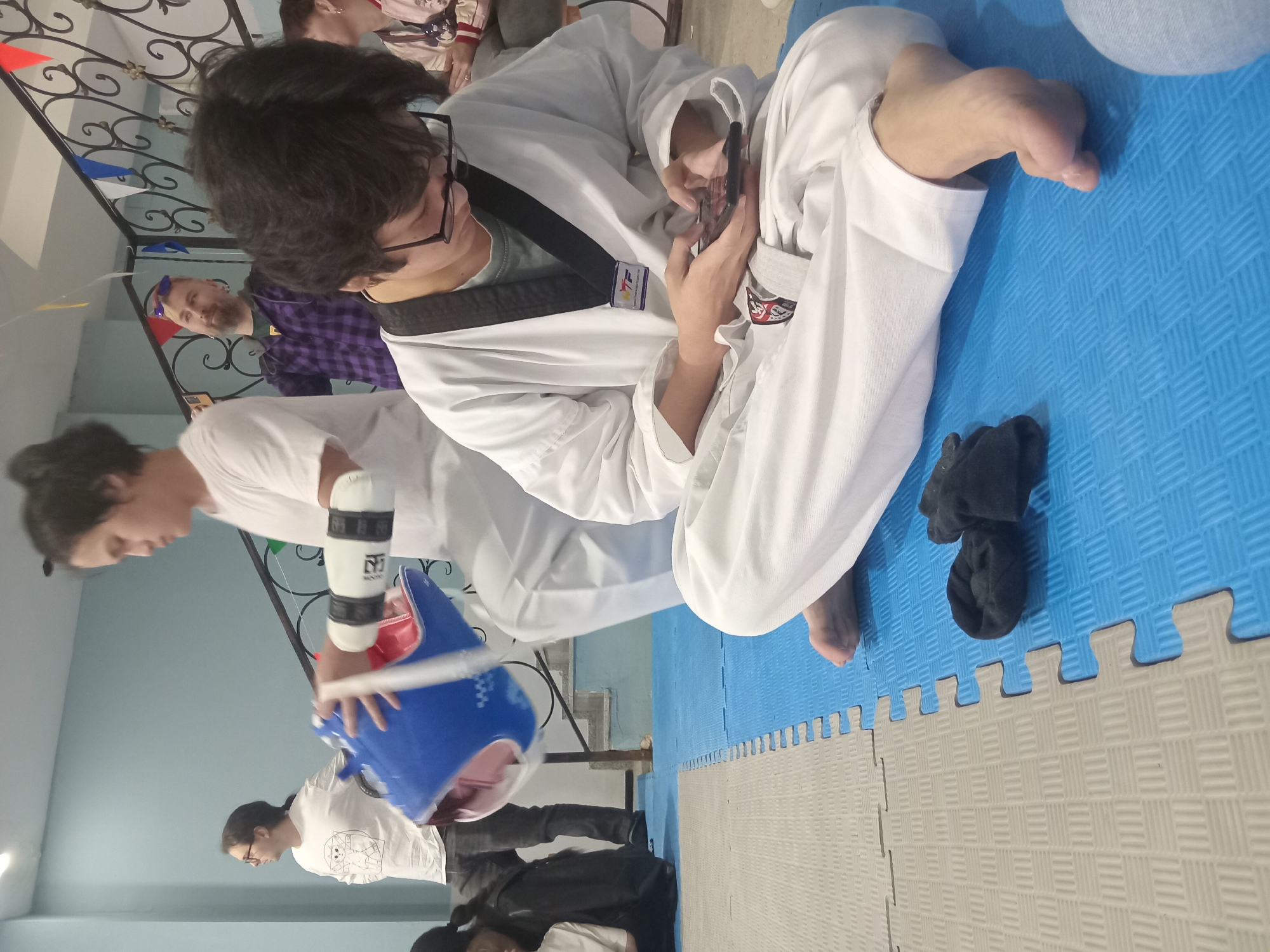 The image shows an indoor scene. A person wearing a white taekwondo uniform with a black belt sits cross-legged on a blue and grey interlocking foam mat and looks at a phone. Black socks sit beside them on the mat. Another person in a white shirt is carrying a blue and red chest protector. A man wearing a purple flannel shirt is visible in the background. Decorative pennants hang from the railing. The location is likely a taekwondo studio or training space. There is no information in the provided image to suggest the location in the city of Batumi, Georgia.