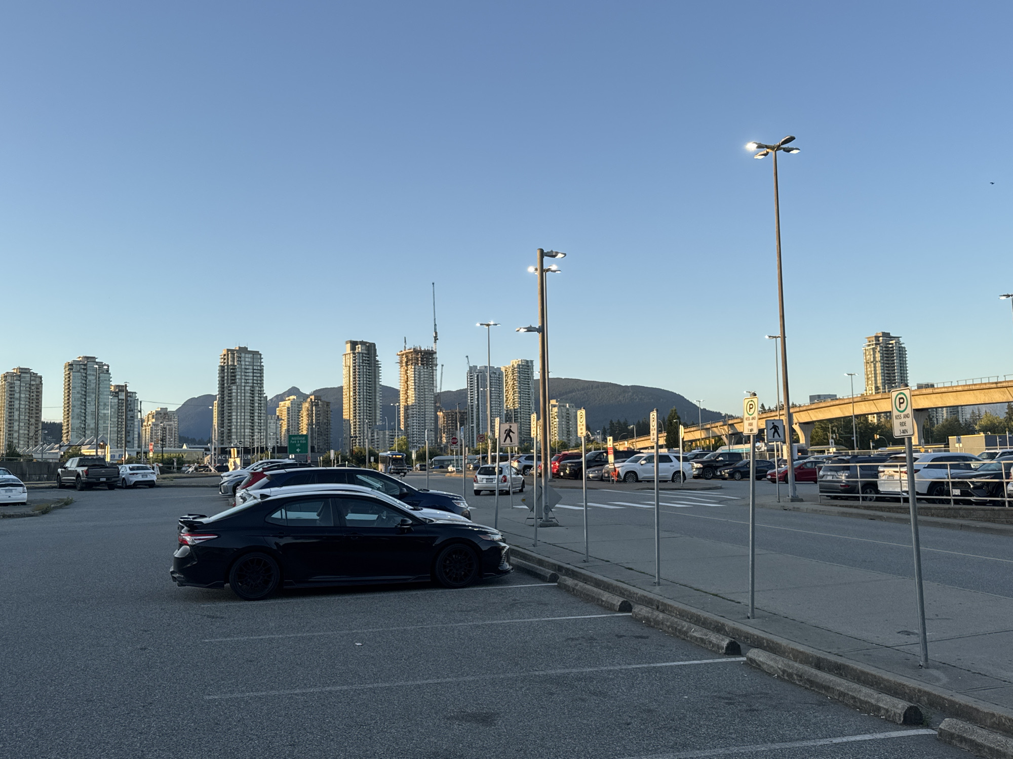 The image captures a cityscape in Coquitlam, Canada, with a focus on a parking lot in the foreground. The parking lot contains several cars, including a black sedan prominently parked on the left. In the background, a line of modern high-rise buildings stretches across the horizon, interspersed with construction cranes, indicating ongoing urban development. The sky is clear and blue, with the sun casting a warm light on the buildings. Several lampposts are visible throughout the parking lot, and traffic signs, including parking restrictions, are present. The overall scene depicts a typical urban environment with a blend of residential and commercial structures.