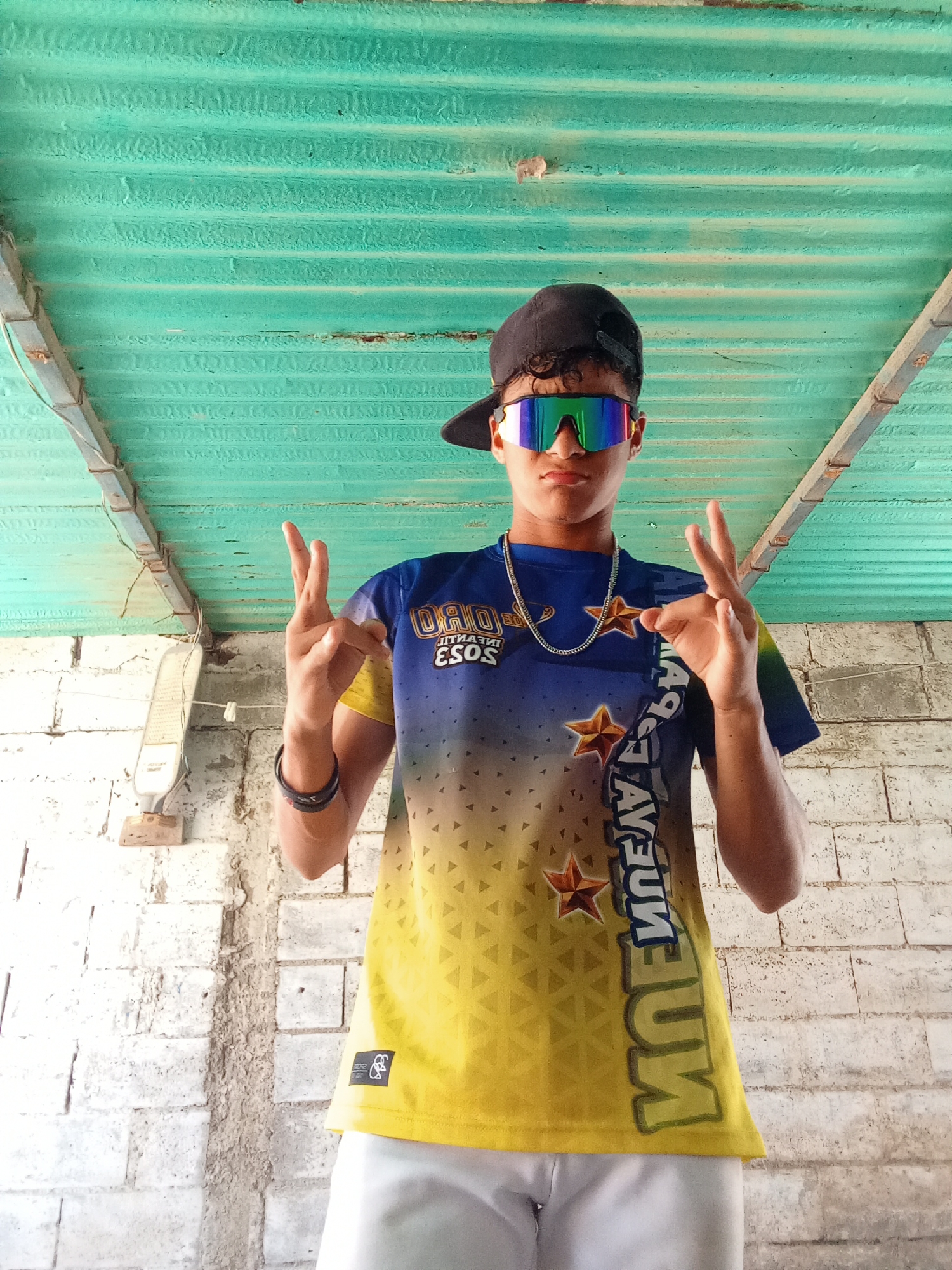 A low-angle, medium shot depicts a teenage male under a corrugated metal roof. The location is Juan Griego, Venezuela. The teen wears a blue and yellow t-shirt with a backwards mirrored word on the right side. He is also wearing white shorts, a black cap with the brim turned backwards, a silver chain necklace, a black bracelet, and iridescent sunglasses. He is making a hand gesture with both hands. The background shows a white brick wall.