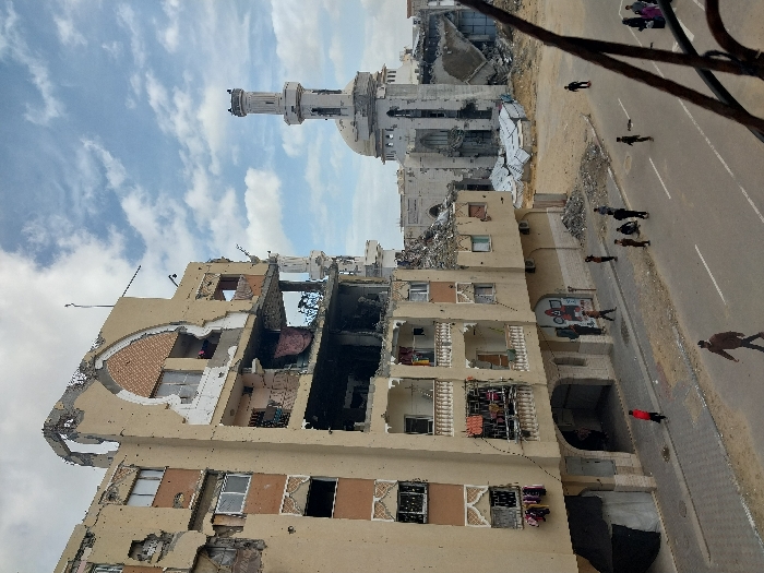 The image depicts a street scene in Khan Yunis, Palestinian Territory, showcasing significant structural damage to multiple buildings under a cloudy sky.
In the foreground, a multi-story building, approximately six floors tall, with a light brown facade, shows extensive damage. The upper floors, particularly the fifth and sixth, have large sections of their exterior walls missing, revealing interior spaces, exposed rebar, and debris. An arched decorative element on the upper facade is partially destroyed. Balconies with white railings are present on the lower levels, with some clothing hanging from them and visible within the damaged interiors. Bullet holes or shrapnel impacts are visible across the building's facade. On the ground level, a recessed archway provides access to the building, and a black tarp structure is set up to the left of the entrance.
To the right and behind this building, a large mosque with a prominent minaret is heavily damaged. The mosque's main structure appears partially collapsed, with its dome showing visible damage and large sections of its walls missing or reduced to rubble. The minaret, though still standing, exhibits damage and erosion, with parts of its upper section fragmented. Surrounding the mosque are piles of debris and rubble.
The foreground features a paved street with white lane markings. Approximately fifteen individuals are present on the street and sidewalk. Several adults and children are walking on the sidewalk and road, while a group of children or young individuals are gathered, some sitting, on the sidewalk near the damaged buildings. One person in a red shirt walks across the street. The overall environment suggests a state of conflict aftermath, with damaged infrastructure and ongoing human presence.