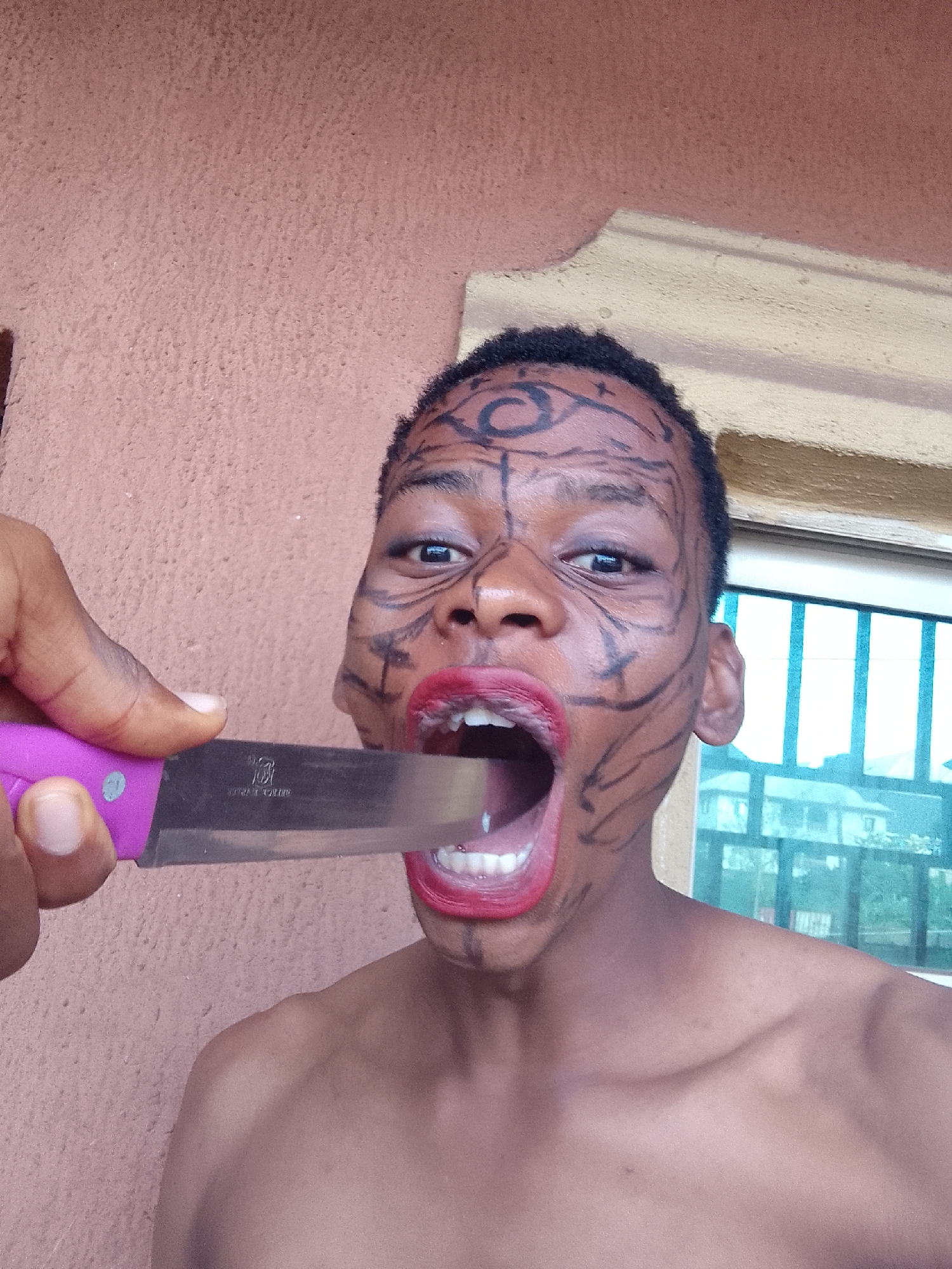 A young person with painted facial designs and bright red lipstick is positioned facing the camera, with a knife partially inserted into their open mouth. The knife has a purple handle and a reflective silver blade. The subject's bare shoulders are visible. The background shows a textured wall of a peach color and a window with a green-tinted view, possibly a residential area with buildings and trees. The location is described as "Unknown, Unknown."