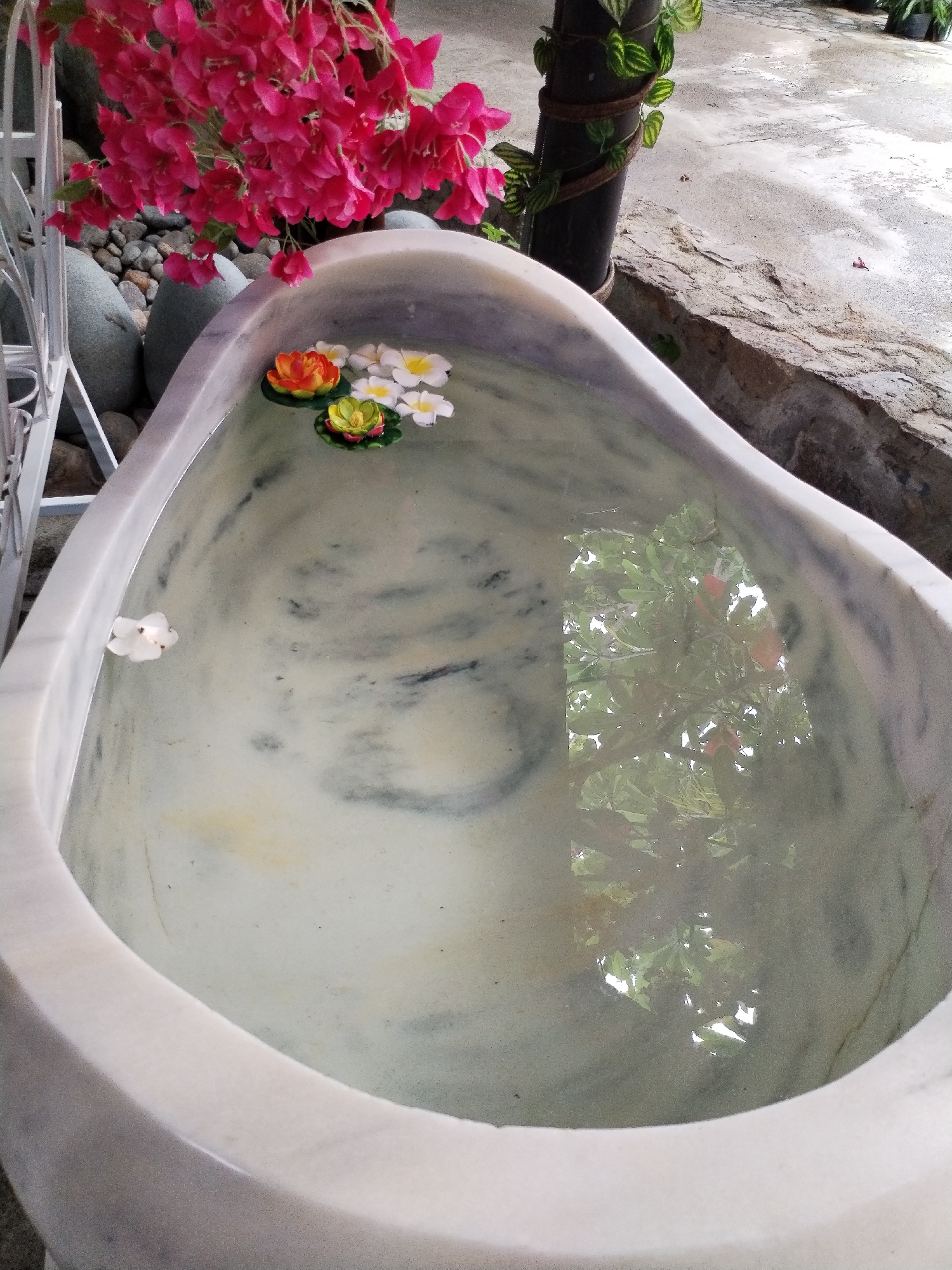 A marble tub-like structure is filled with water, reflecting foliage above. Floating on the water's surface are several artificial flowers in shades of orange, yellow, and white. A single white flower petal floats on the left. A pink flowering plant drapes over the side of the tub. The tub is positioned next to a stone path and decorative elements, including a metal frame and rocks. A plant-wrapped pole stands in the background. The scene appears to be located in a garden or outdoor decorative space in Nam Nha Trang Ward, Vietnam.