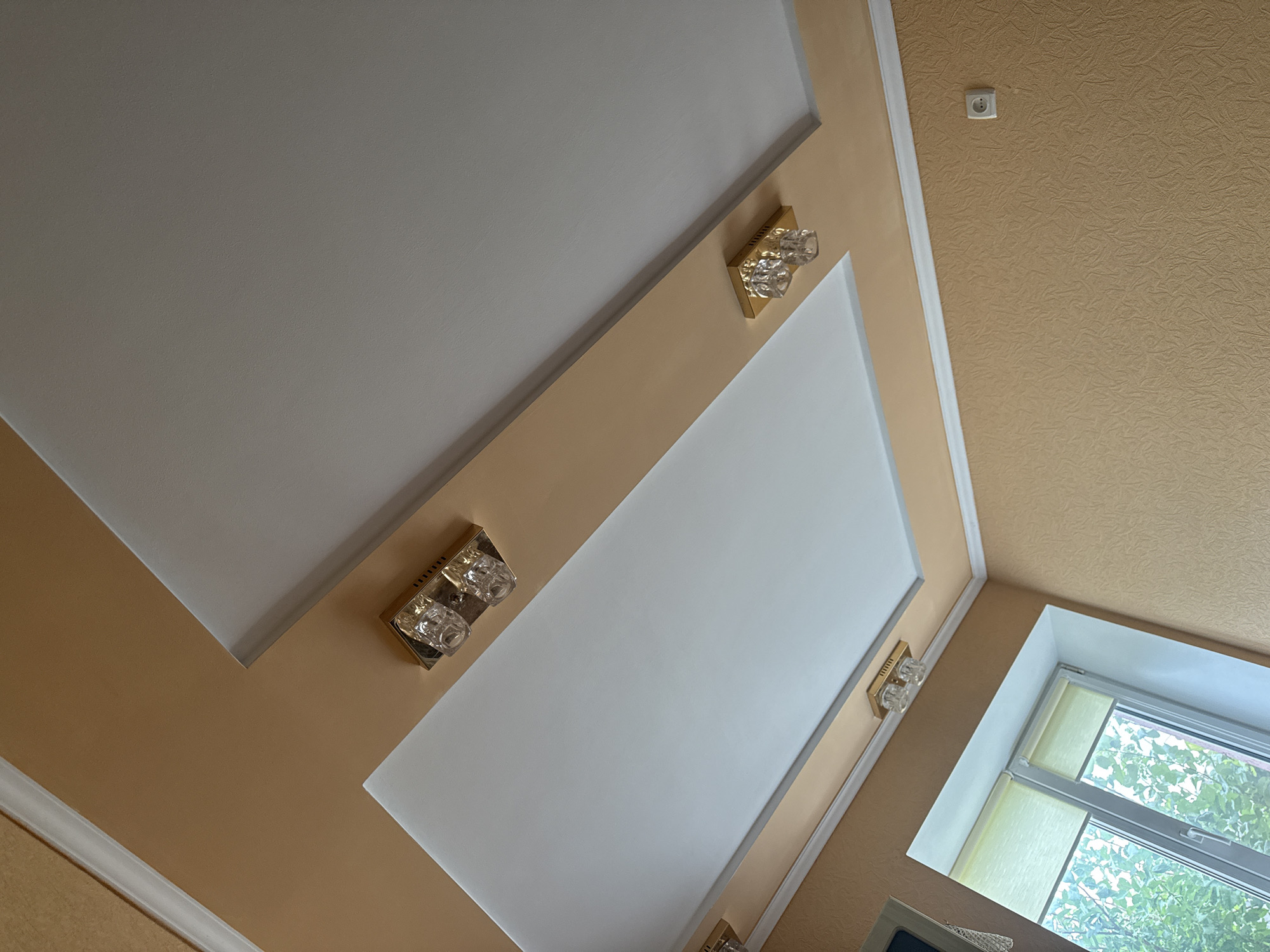 A multi-level ceiling painted in white and beige is visible in this indoor shot. Two identical wall-mounted light fixtures with crystal shades and gold bases are attached to the beige sections of the ceiling. One is located in the upper left and the other in the lower right portion of the image. A white window frame with green blinds is visible in the lower left corner. The walls are painted beige and textured.