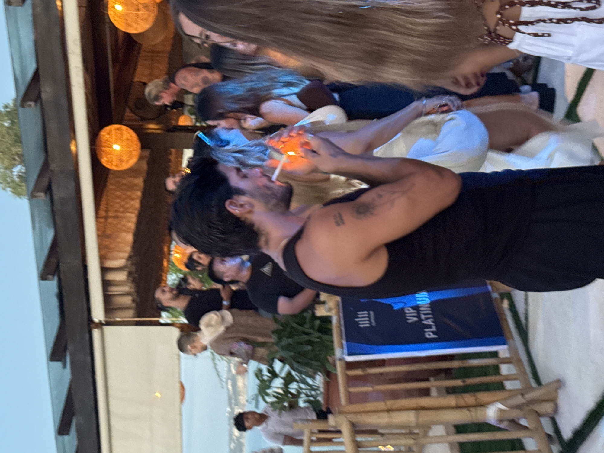 This image depicts an evening social gathering at an outdoor or semi-outdoor venue, likely a bar or restaurant, in Kuta Selatan, Indonesia. The scene is illuminated by warm, ambient light from several round, woven pendant lamps, suggesting dusk or early evening.
In the foreground, a man with dark hair and a beard, wearing a black tank top, is actively lighting a cigarette with a lighter. The flame clearly illuminates his hand and the cigarette. A tattoo is visible on his right bicep. His arm is around another individual, with long blonde hair, whose face is also subtly lit by the flame.
The midground and background feature several other people, some standing and others seated, appearing to socialize in a relaxed setting. The venue incorporates natural materials, including visible wooden beams and bamboo furniture. On the right side of the image, a blue sign is partially visible, bearing white text that clearly reads "VIP PLATINUM" along with a logo resembling "1111". In the distant background, a hazy view of a landscape, possibly a coastal area with water, suggests an elevated location. Some green foliage is also present, enhancing the natural and relaxed ambiance of the gathering.