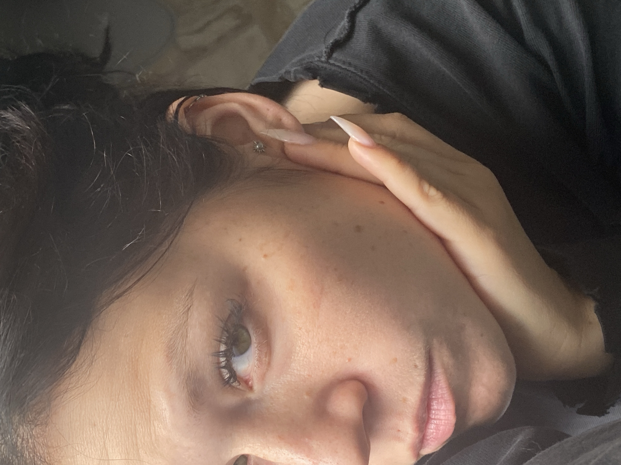 A close-up, profile view shows a young Caucasian woman with dark hair pulled back, resting her chin on her right hand. Her left eye, a light green color, looks directly at the camera. She has a fair complexion with scattered freckles and faint lines on her forehead. Her lips are a natural pink hue, and her eyebrows are dark and lightly shaped. She is wearing a dark, hooded garment. Her fingernails are long and light pink, with the index finger of her right hand touching her cheek near her ear. A small, silver stud earring with a floral design is visible in her right earlobe, and a thin silver hoop earring is in her upper ear. The background is softly lit and out of focus, suggesting an indoor setting. No specific location in Chaikovsky, Russia, can be determined from the visual information.