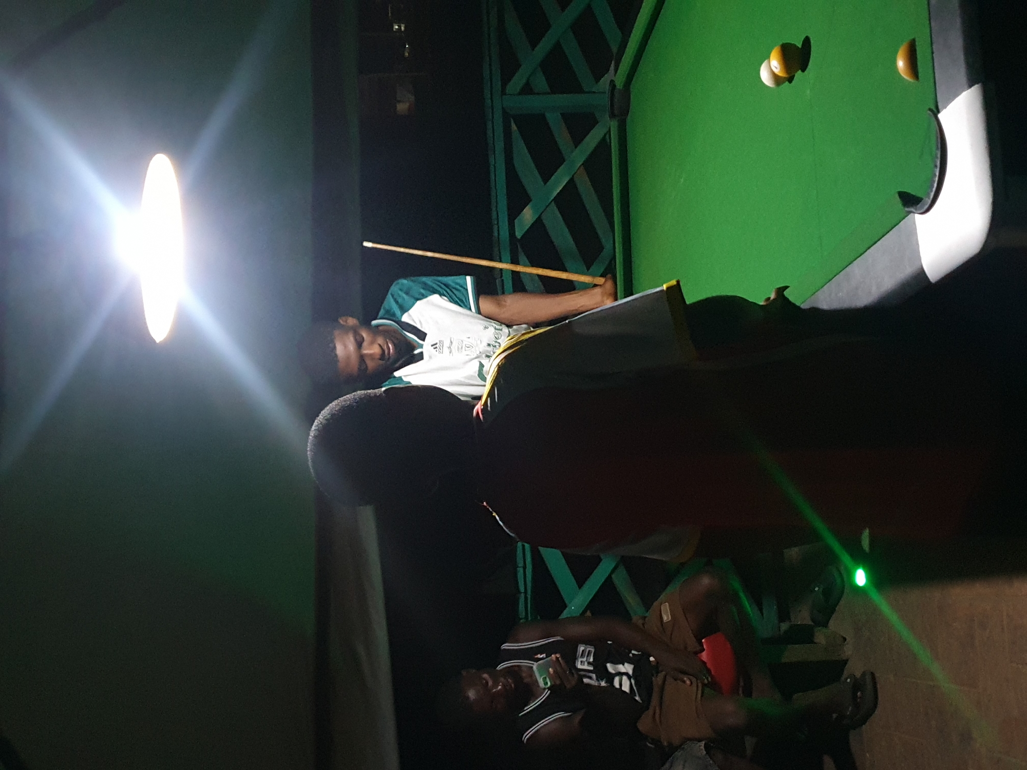 The image depicts an outdoor billiards area at night, likely in Lokoja, Nigeria, illuminated by a bright overhead light. In the foreground, a person is seen from behind, facing the pool table. To the right, another person, wearing a green and white shirt, stands over the pool table, holding a cue stick, appearing to play a game. Three billiard balls are visible on the green felt surface of the table: one white, one yellow, and one partially obscured orange ball near the pocket. In the background, another individual is seated, looking down at a mobile phone. The environment includes a green railing structure behind the pool table and a dark, possibly covered, ceiling overhead. The overall scene suggests a casual recreational gathering.