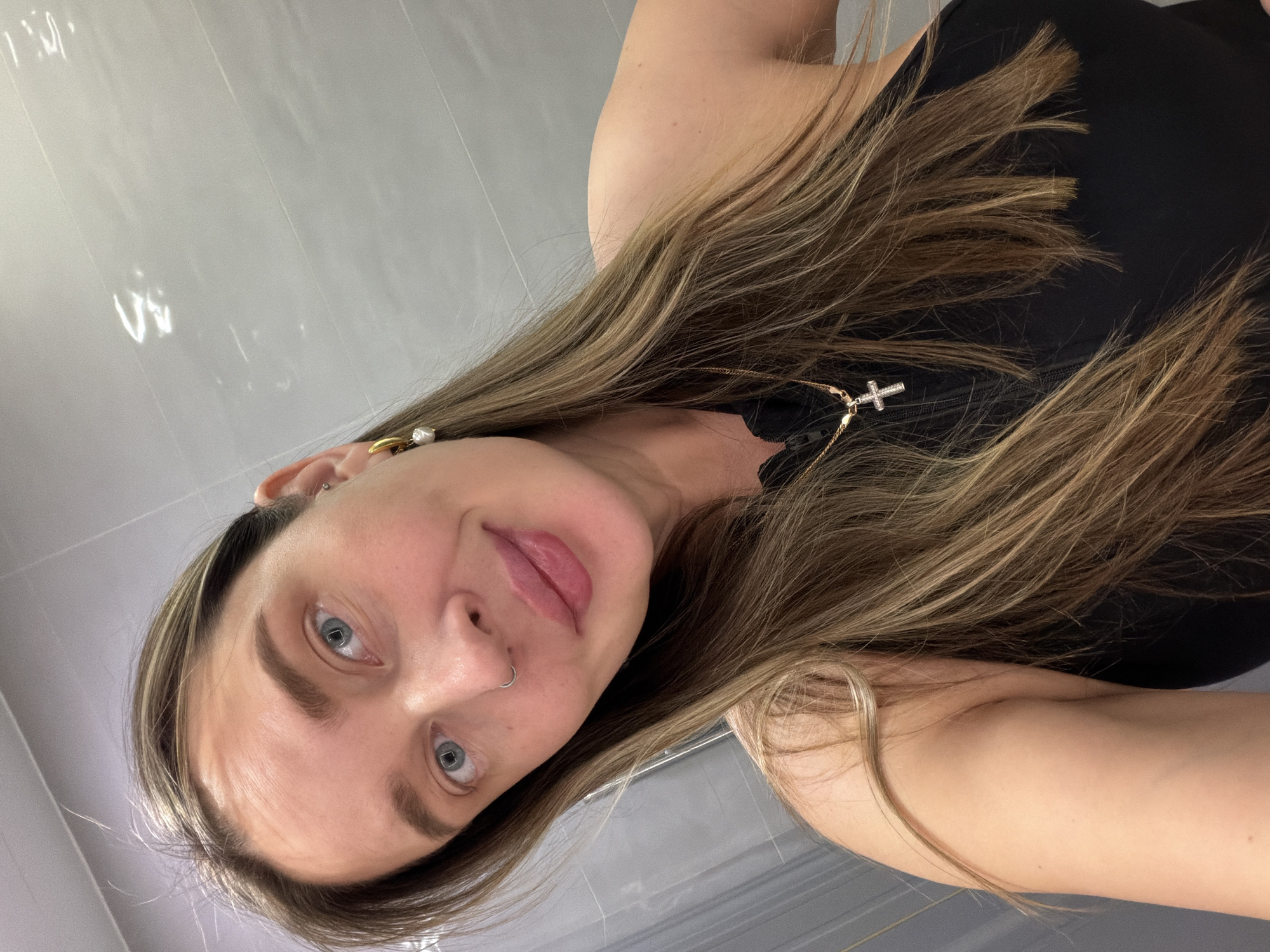 A woman is posing for a selfie in a bathroom. She has light brown hair with blonde highlights, blue eyes, a nose ring, and is wearing a black top. She is wearing a cross necklace. The bathroom walls appear to be tiled in gray. It is not possible to verify the location of the image as Khimki, Russia.