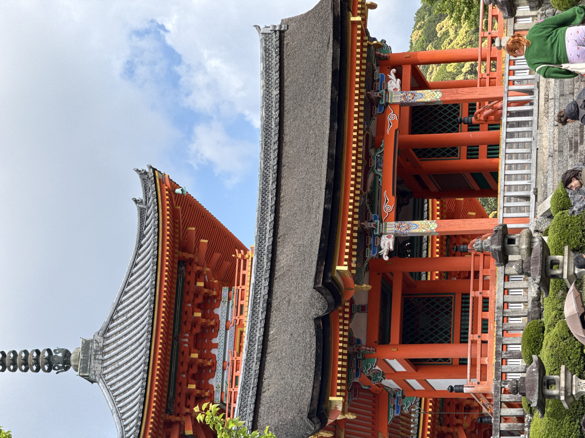 This image captures a vibrant scene at a traditional Japanese temple, likely in Kyoto. The main focus is on the intricate architecture of the temple buildings, characterized by striking orange-red wooden pillars and beams. These structures are adorned with gold accents and colorful decorative carvings, including distinct white swan-like figures and ornate patterns.
Two main temple roofs are visible: one in the foreground on the right has a dark, textured surface, possibly thatched, while the other, further back on the left, features a lighter, multi-layered tiled roof culminating in a decorative finial at its peak. The architecture stands against an overcast sky with hints of blue, and lush green, possibly mountainous, terrain in the background.
In the foreground and mid-ground on the right, several visitors are present, suggesting the temple is a popular cultural or tourist destination. A woman with reddish-brown hair, dressed in a green sweater and patterned pants, is visible looking downwards. Another person with dark hair is partially seen in the lower right, and a beige umbrella is held by a third, partially obscured individual. These people appear to be exploring the temple grounds.
Adding to the serene atmosphere are manicured green bushes and two ornate stone lanterns. Japanese characters are visible on one of the stone lanterns, providing a specific cultural cue. The image depicts a daytime scene, likely on a cloudy day, highlighting the temple's detailed craftsmanship and colorful elements amidst a natural setting.