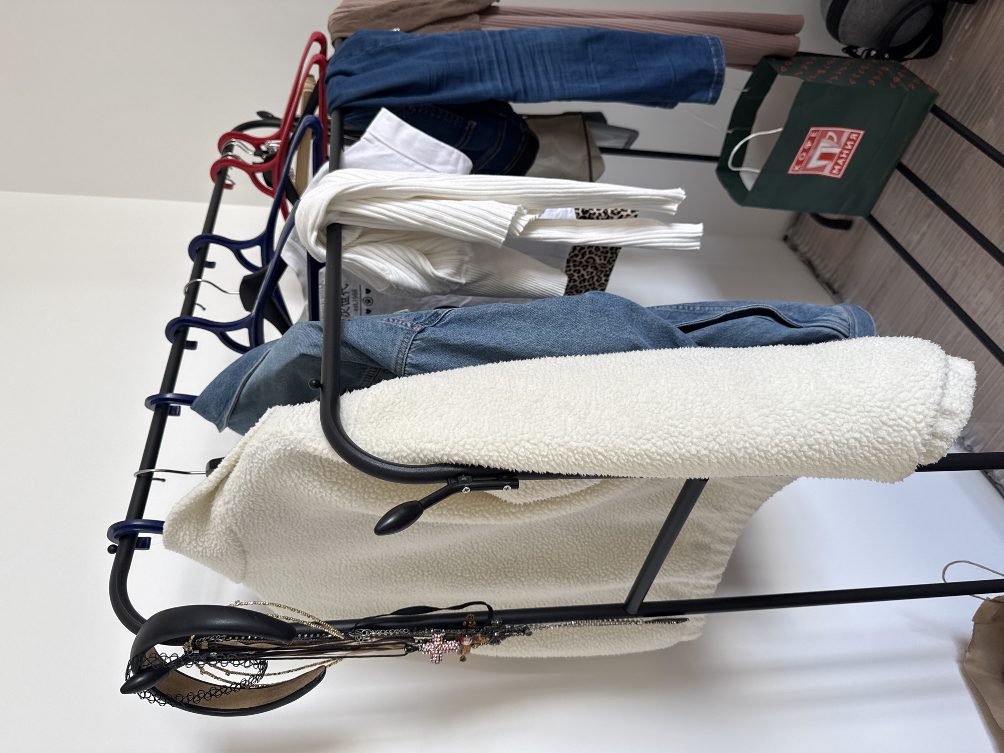 The image shows a black metal clothes rack against a white wall, leaning slightly to the right. Clothes and accessories are draped over the rack. At the top of the rack are items of clothing, including what appears to be blue denim and a light-colored ribbed knit top. A green tote bag with a white handle and a red logo reading "КОФЕМАНИЯ" is hanging on the right. This translates to "Coffee Mania," a chain of coffee shops with locations in Moscow, Russia. Further down the rack, there's more denim and a fluffy white jacket or blanket. At the bottom, a black belt with a lacy edge and assorted jewelry are visible. Hangers in various colors, including red and blue, support some of the clothing items. The background includes a textured surface, possibly a door or wall with vertical stripes.