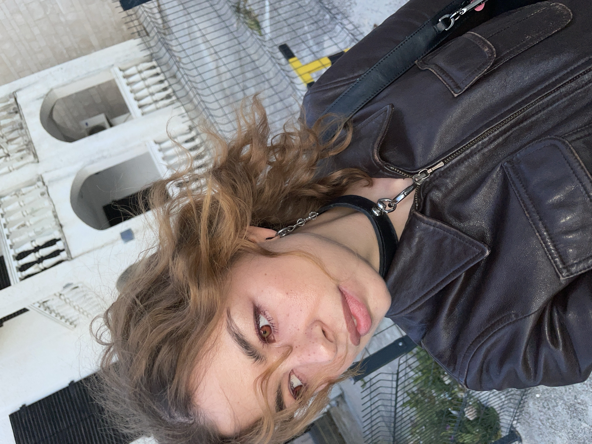 The image presents a close-up, rotated portrait of a young individual, likely female, with light brown, curly hair. She looks upwards towards the camera with a neutral expression, her lips slightly parted, revealing warm brown eyes and freckles across her nose and cheeks. She is wearing a dark brown, possibly distressed, leather jacket with visible zippers and pockets. A black choker-style necklace with a silver chain and ring adorns her neck, and a black strap, likely from a bag, is seen over her shoulder.
The background suggests an outdoor, elevated setting, possibly a balcony or terrace, looking down onto a scene. Below, there are white or light-colored buildings featuring distinctive arched window openings and horizontal slatted structures, which could be railings or shutters, characteristic of Mediterranean architecture. A dark, grid-like fence or railing is visible in the foreground and mid-ground. Green foliage is also present, along with a glimpse of a bright yellow object in the distance. The lighting indicates it is daytime with clear weather.
The architectural style, featuring white buildings with arches, aligns with coastal regions like Boreti, Montenegro, providing a specific location cue. No visible text is present in the image.