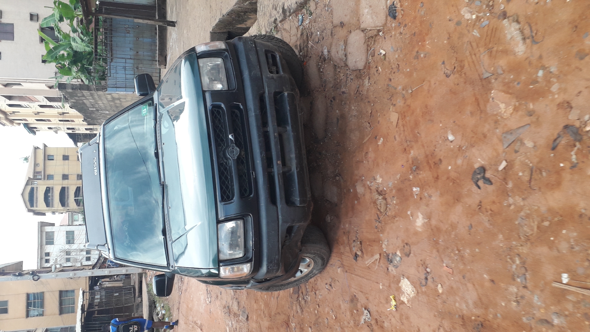 The image depicts a green Nissan SUV parked on a dirt road in what appears to be a residential area in Onitsha, Nigeria. The ground is littered with debris. Buildings are visible in the background. A man in a blue shirt stands near the edge of the frame.