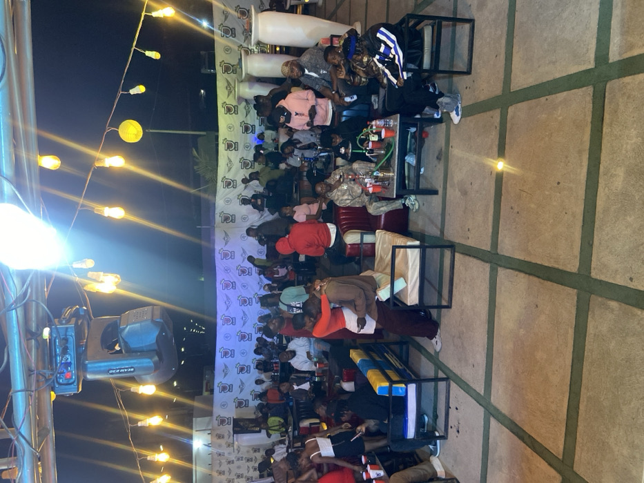 A group of people sit at tables in an outdoor setting, enjoying beverages and conversation. A large banner is visible in the background, and string lights hang overhead, creating a festive ambiance. Some individuals are seated on cushioned benches. A stage lighting fixture is visible on the left of the frame. The ground is paved with large, square concrete slabs.