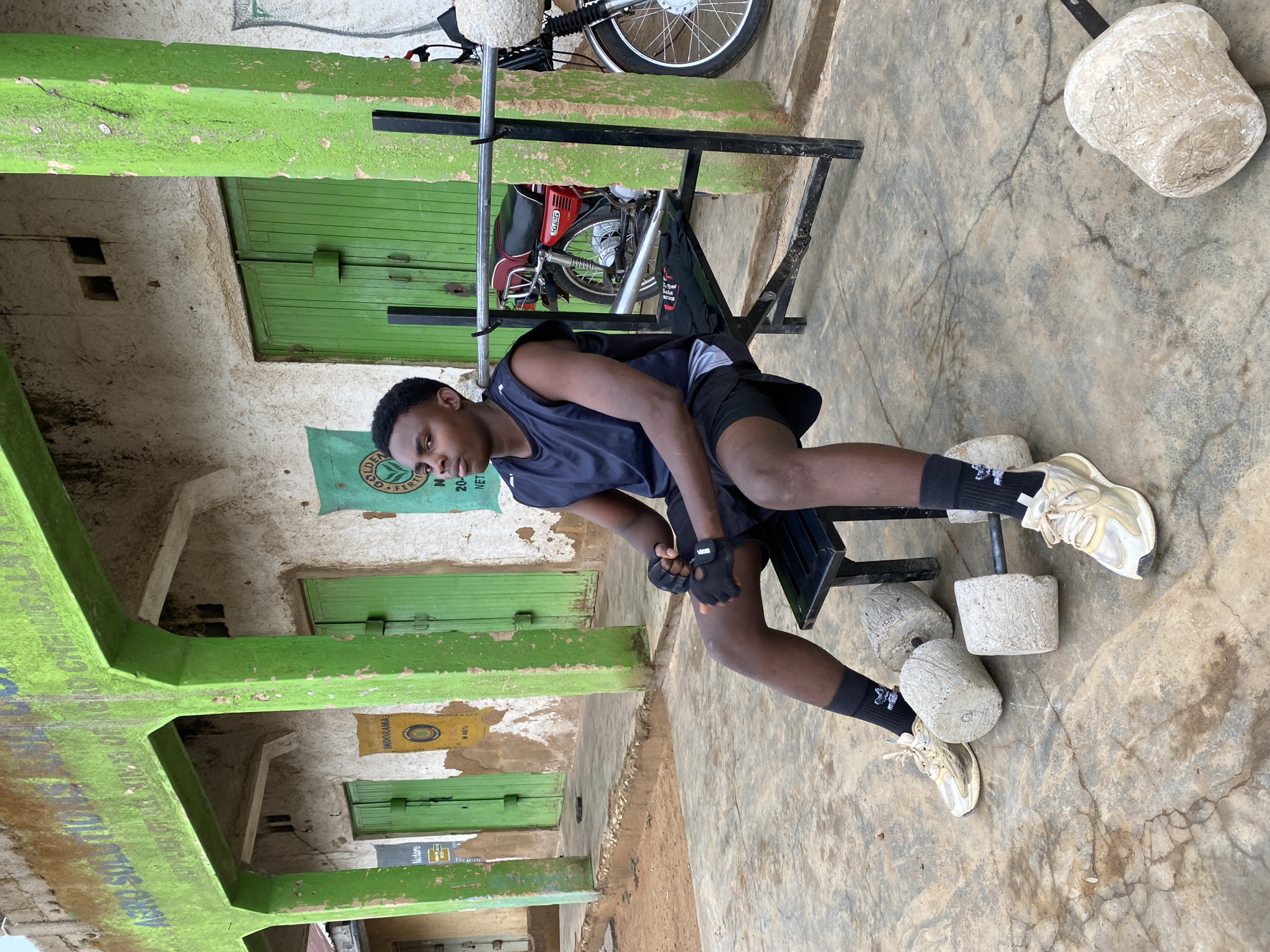 A young man is performing a leg exercise, likely seated leg extensions, in an outdoor setting in Riyom, Nigeria. He is wearing athletic clothing: a sleeveless dark blue shirt, black shorts, black socks, gloves, and off-white sneakers. He sits on a black metal weight bench, with homemade concrete weights attached to the leg extension bar. A red motorcycle is parked behind the weight bench. The setting appears to be in front of a building with weathered green-painted columns and doors. Advertising signs or posters are visible on the building's facade. The ground is concrete and shows signs of wear and cracking.