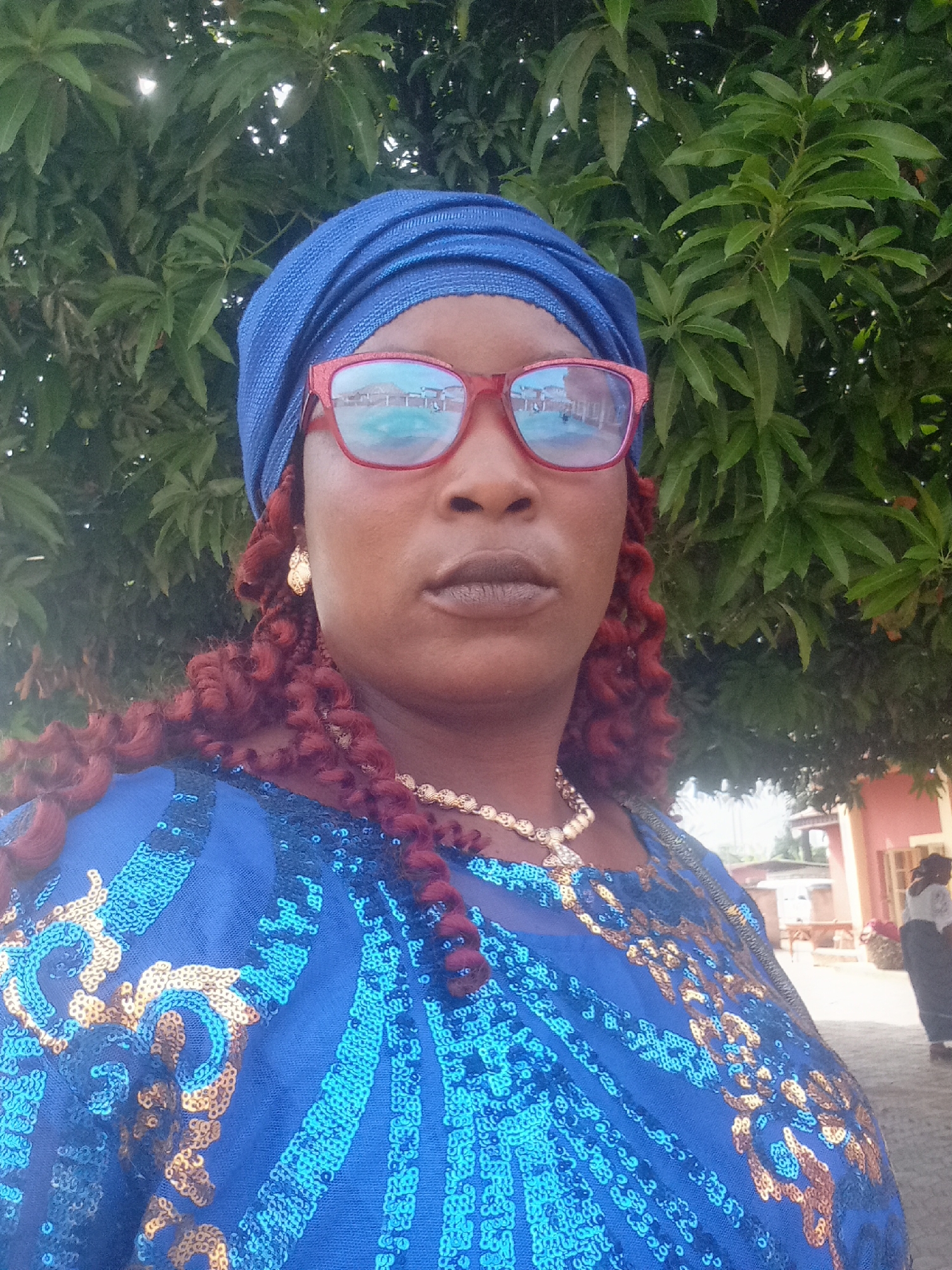 A medium shot depicts a woman outdoors. She wears a blue headwrap, red-framed eyeglasses, a gold necklace, and earrings. She has brown skin and reddish-brown curly hair. Her blue top is adorned with gold sequins. The background includes green foliage, and a building is visible. Based on the building's design and known location, the picture was taken in the city of Akwanga, Nigeria.