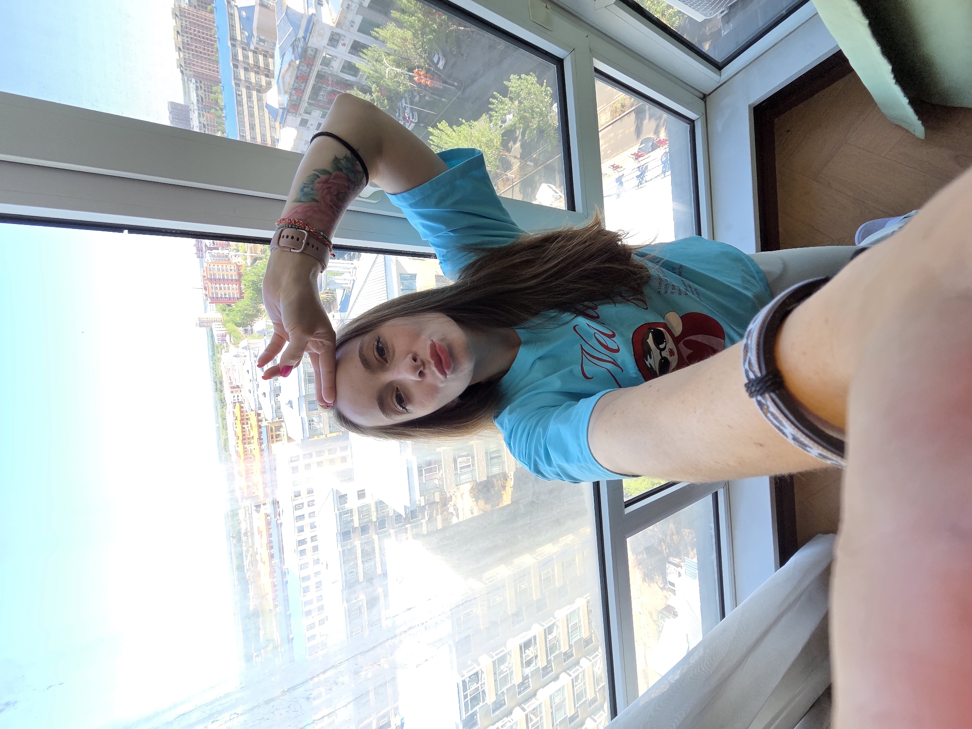 A young woman is captured in a selfie taken indoors, likely in a high-rise building with a panoramic city view. She has fair skin, long brown hair, and dark eyes, and she is looking directly at the camera with a slightly pursed-lip expression. Her left hand is raised towards her forehead, fingers slightly curled, in a casual pose.
She is wearing a light blue short-sleeved t-shirt adorned with a graphic of a stylized red figure, possibly a Daruma doll or character wearing sunglasses. Text visible on her shirt includes "Hello" and partially legible phrases like "simple with a new look." On her left forearm, a colorful tattoo with a floral design, possibly roses, is visible. Her left wrist features a light pink smartwatch and several beaded bracelets, while a black hair tie is on her upper left arm. Her fingernails are painted a reddish-pink hue.
Behind her, large, white-framed windows reveal a bright, sunny cityscape, indicating it is daytime with clear weather. The view encompasses numerous multi-story buildings and green trees, characteristic of an urban setting such as Tongjiang, China. A light-colored curtain is visible on the right side of the frame, and a wooden floor can be seen in the bottom right corner, suggesting an interior living space. The image is well-lit by natural light from the window.