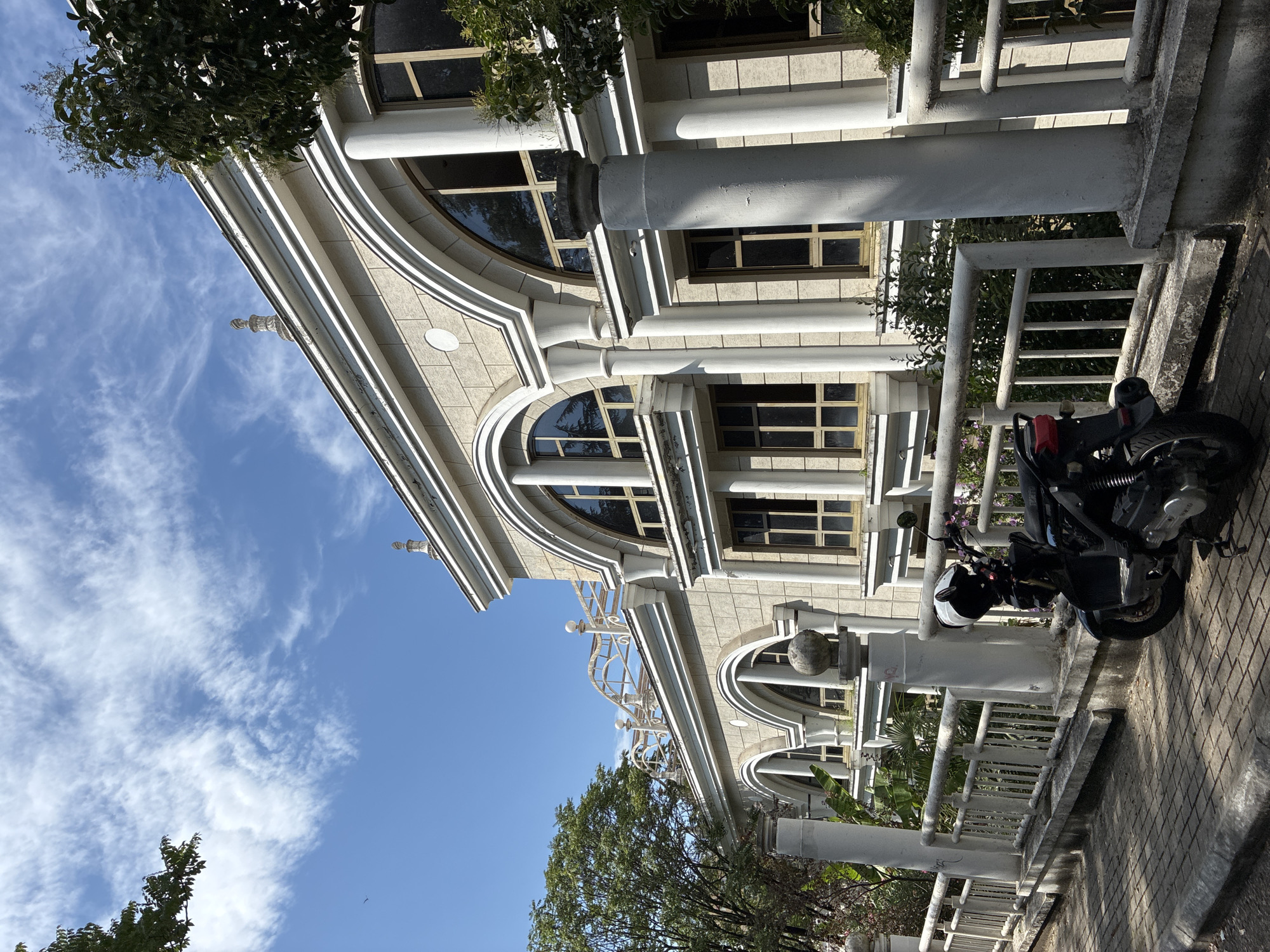 The image shows a building with neoclassical architecture, featuring arched windows and columns, likely located in the city of Sochi, Russia. The building's facade is made of light-colored stone. In the foreground, a parked black scooter with a helmet on its handlebars is visible on a cobblestone path. A white fence runs alongside the path, separating it from a lush green area behind it. The sky is blue with scattered white clouds, and trees with green foliage frame the scene.